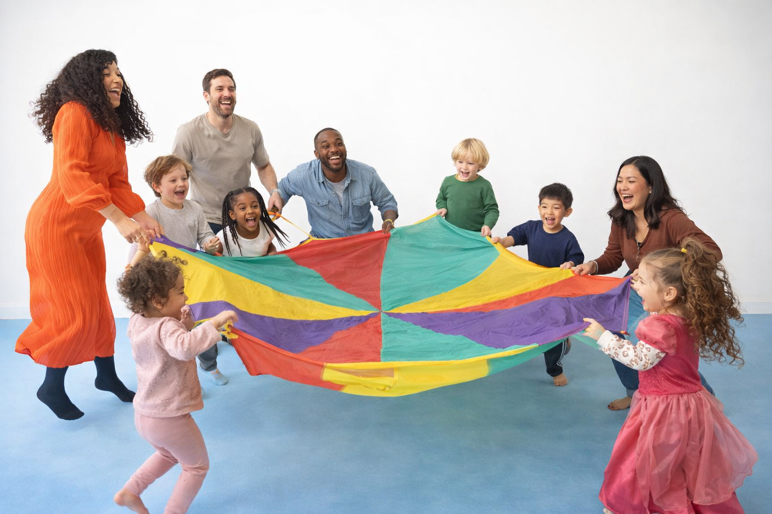Children and adults playing with a colorful parachute in a room with a plain white wall.