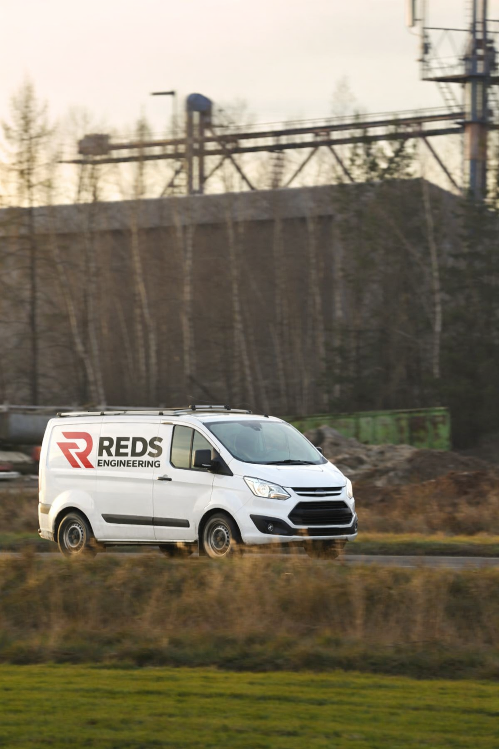 A white van with the logo and text 'REDS Engineering' on its side, driving on a road with a background of trees and industrial structures.