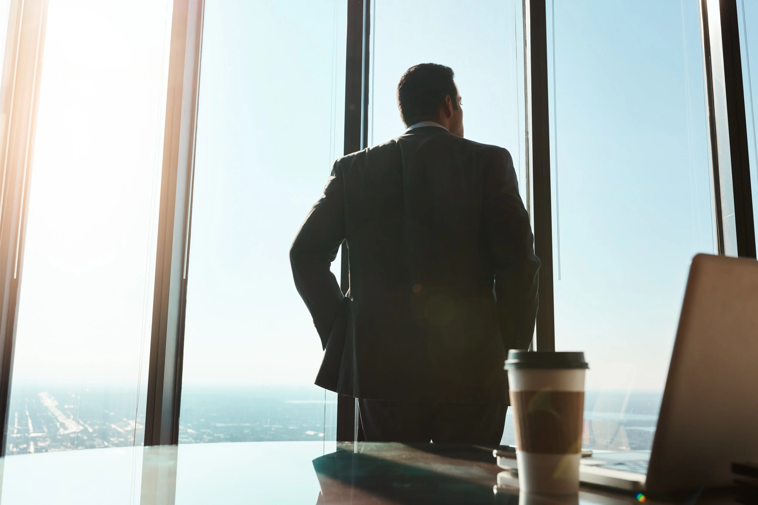 A man in a business suit looking out a large window in a high-rise building, with sunlight streaming in and a city view below.