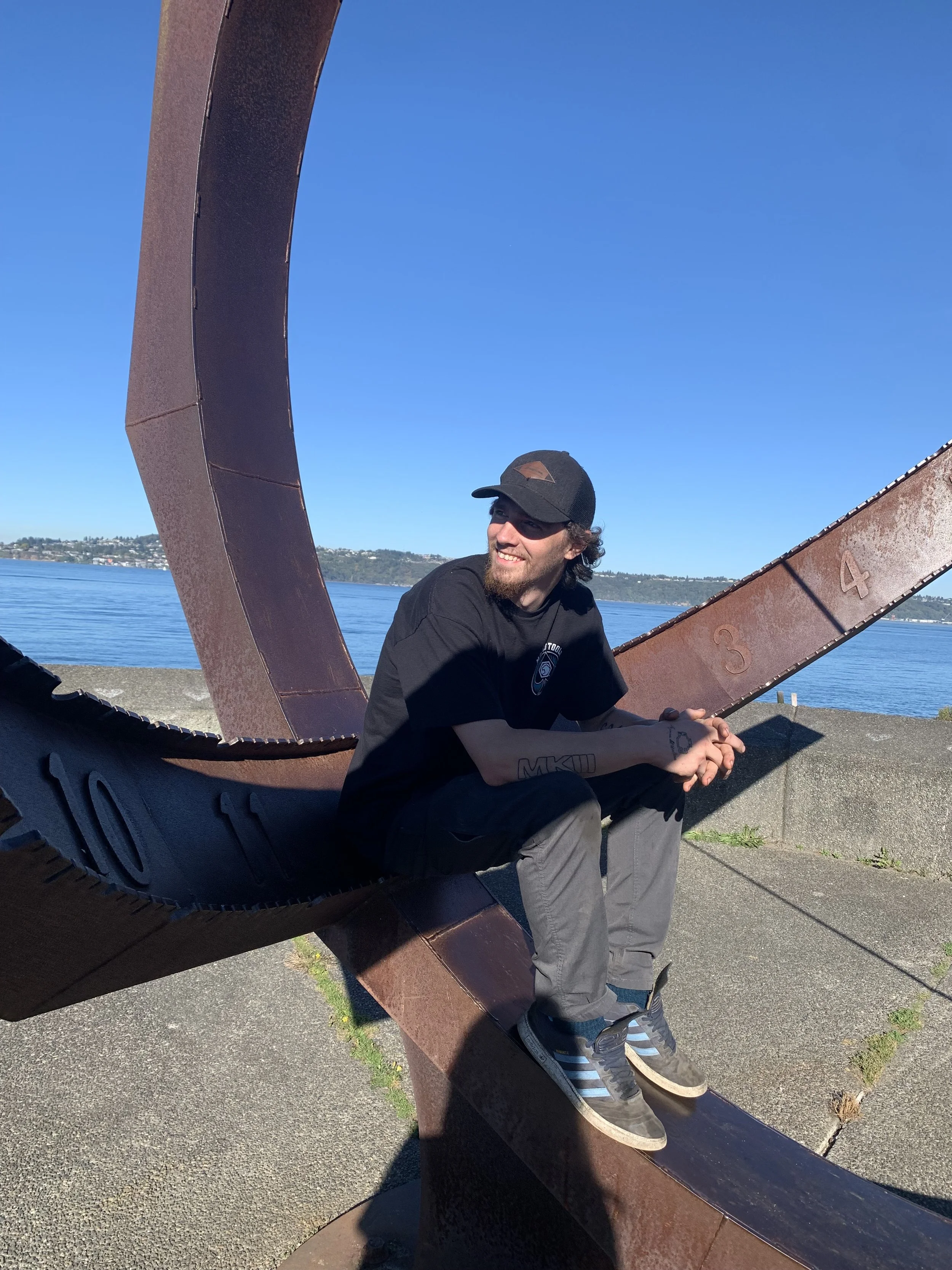 A man with a black cap and black T-shirt sitting on a large metal sculpture by the water, smiling and looking to the side, with clear blue sky and water in the background.