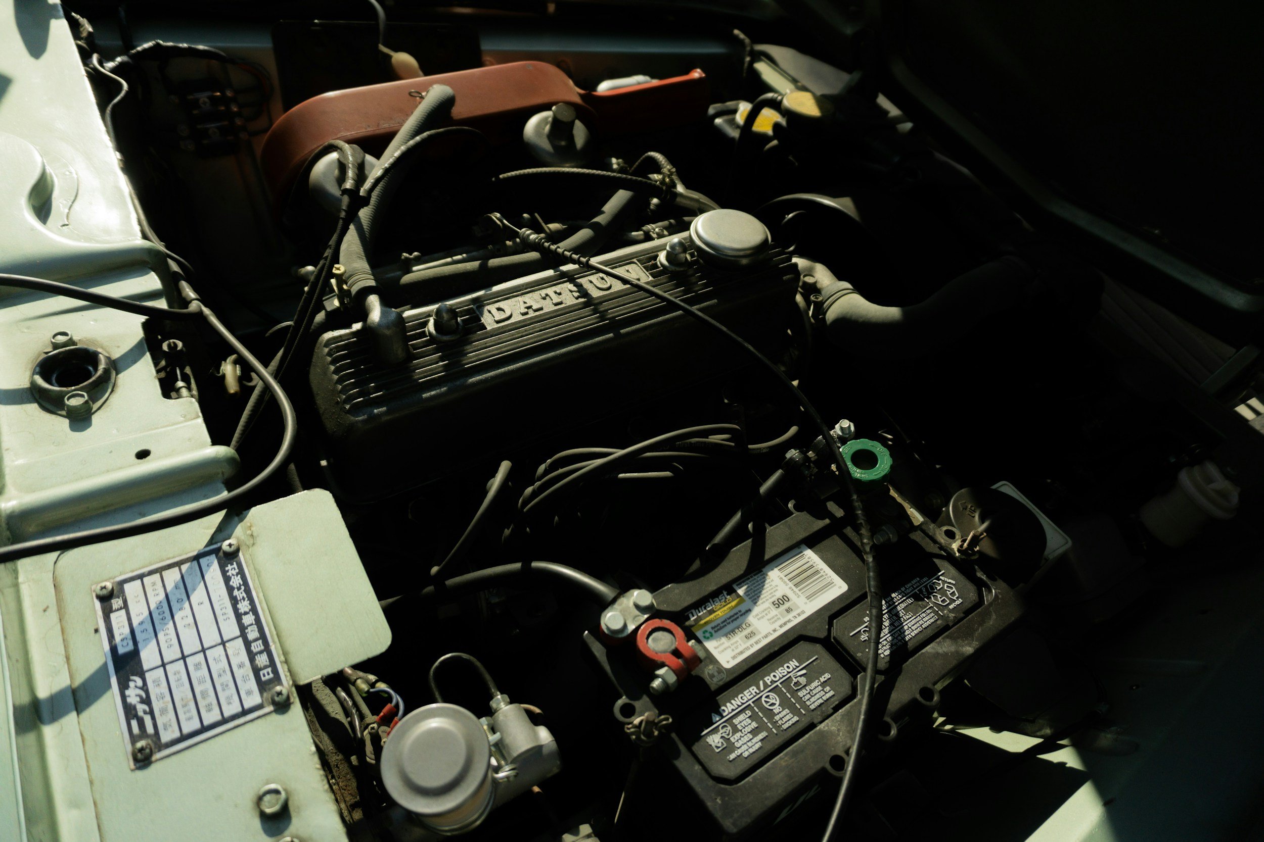 Car engine with various components including a black valve cover marked 'DATSUN' and a car battery with labels, wires, and hoses.