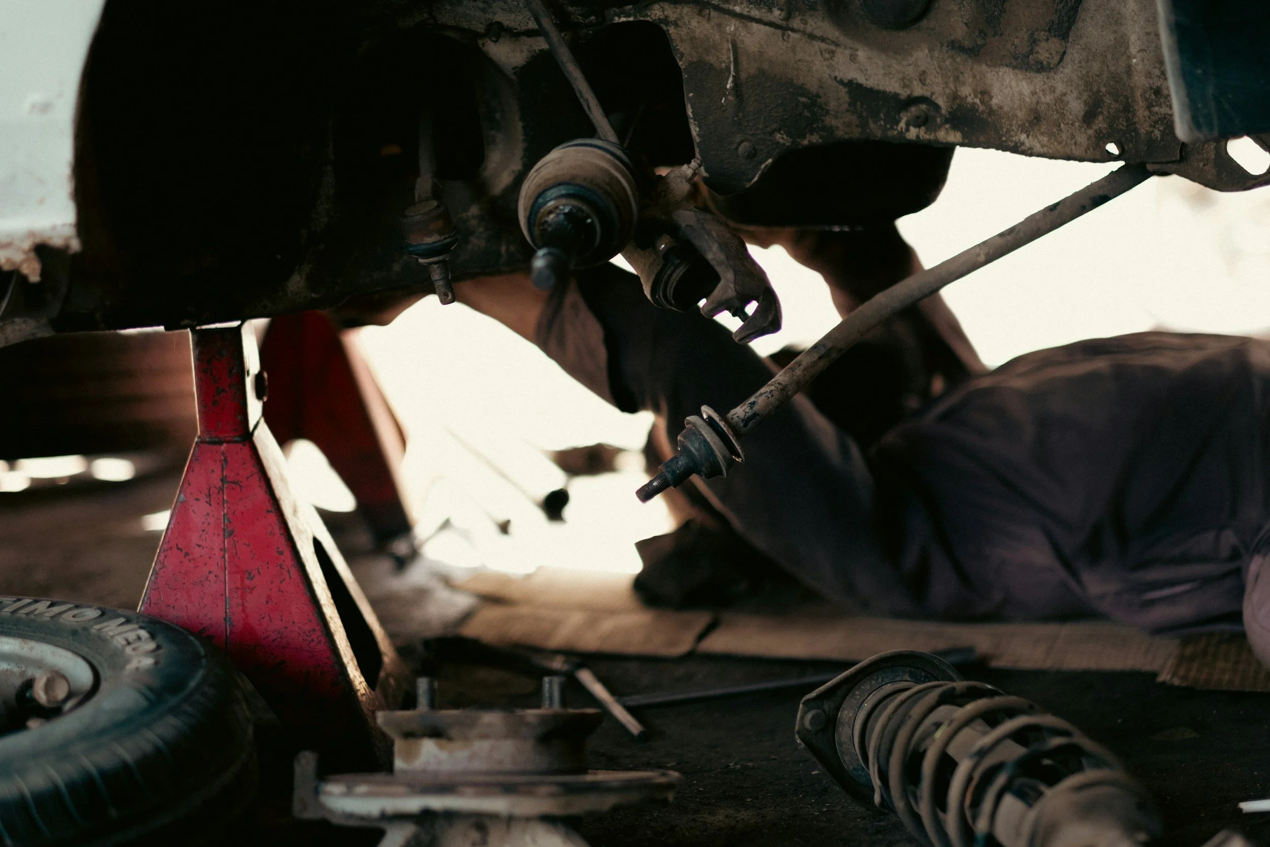 A mechanic working underneath a car, which is supported by a red jack, with tools and car parts around in a garage.