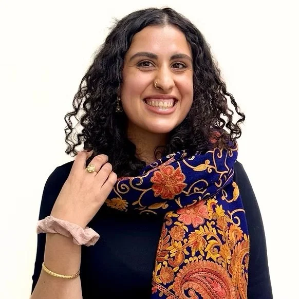 A woman with curly black hair smiling, wearing a black top, colorful embroidered scarf, and gold jewelry, against a plain white background.