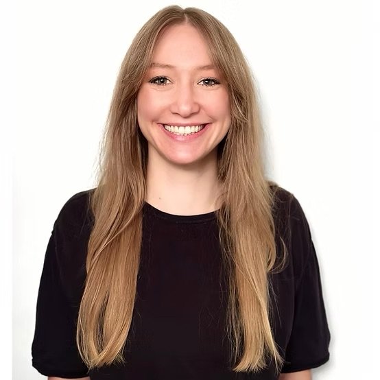 A young woman with long blonde hair smiling, wearing a black shirt, against a plain white background.