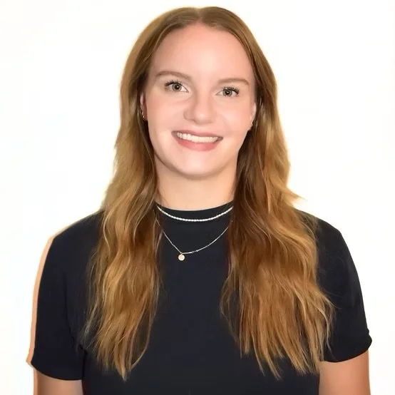 Young woman with red hair smiling, wearing a black top and layered necklaces, against a white background.