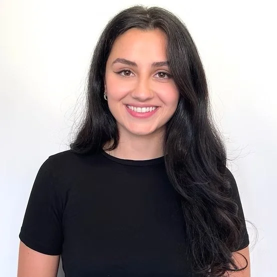 A young woman smiling with long dark hair, wearing a black shirt, standing against a white background.