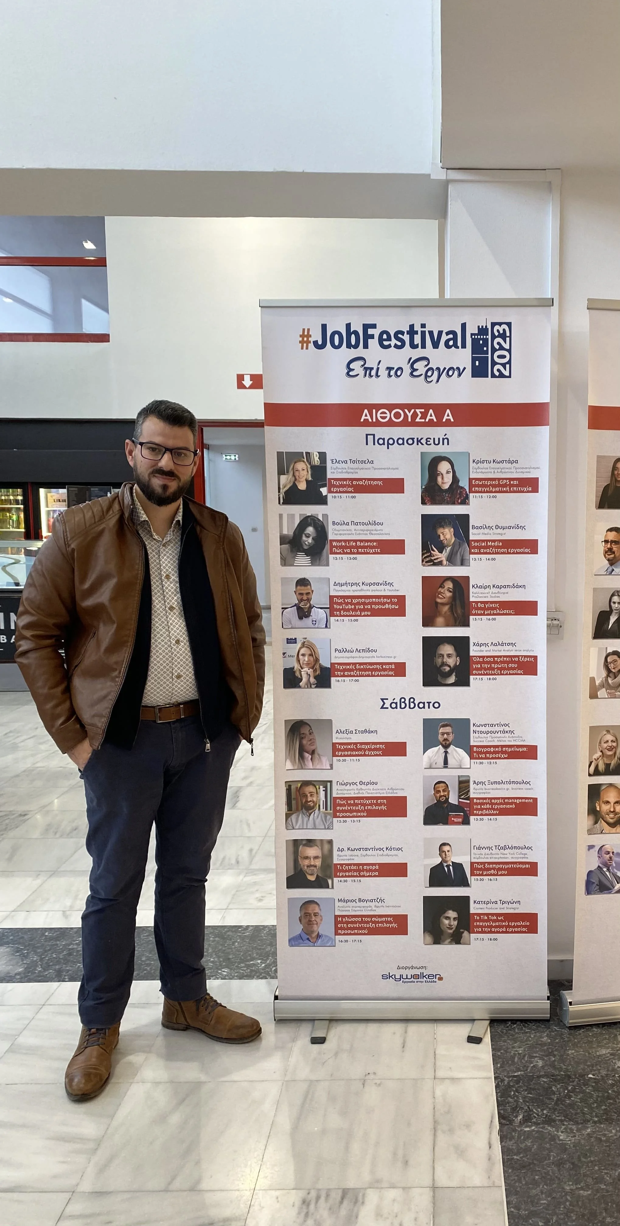 A man standing next to a poster for the #JobFestival 2023, with schedule and speaker details written in Greek, inside a building.