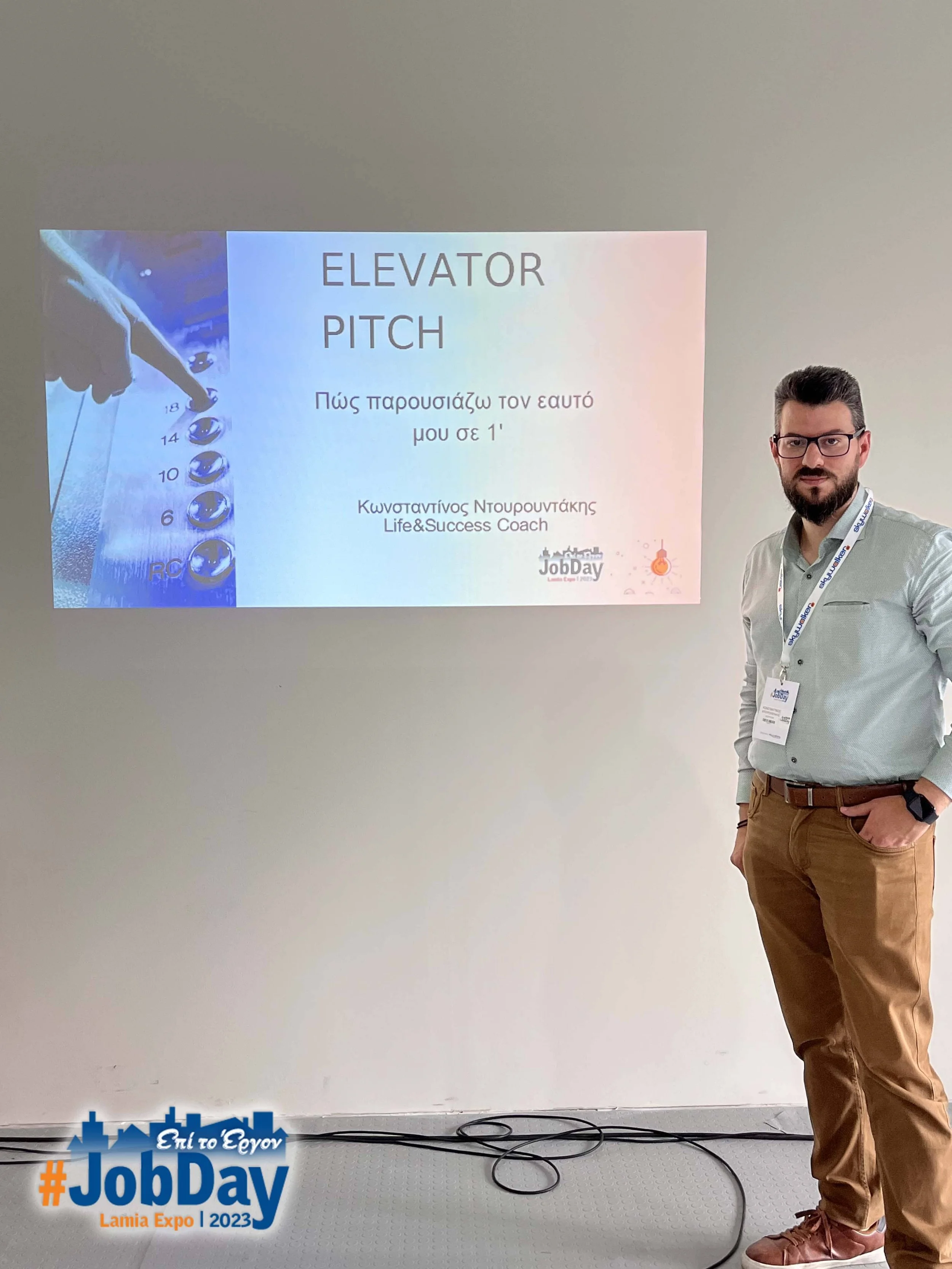 Man standing next to a presentation slide about elevator pitch at a JobDay event in Lamia Expo 2023, with a white wall behind him.