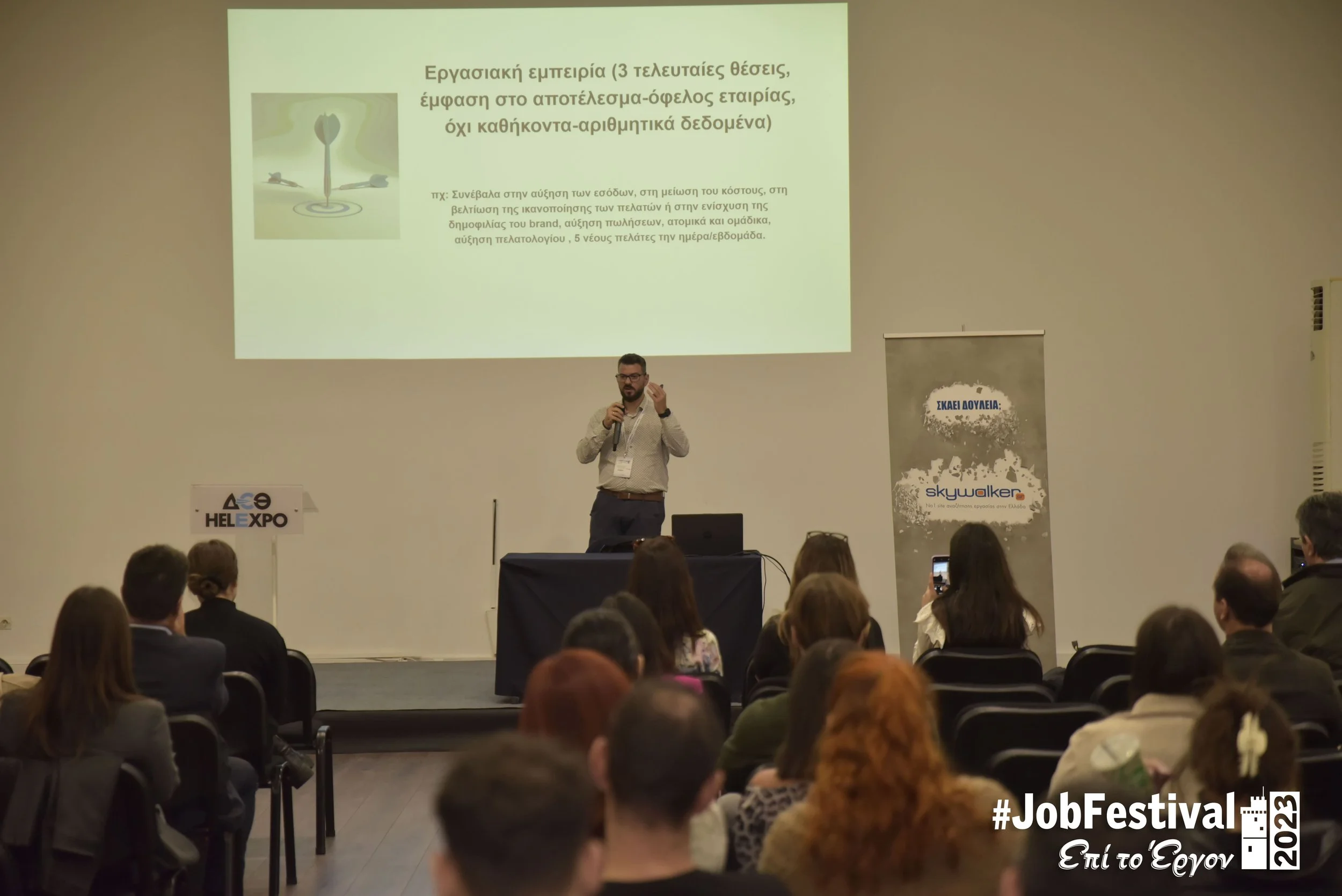 A man giving a presentation on a stage at a conference, with a slideshow projected on the screen behind him. The audience is seated, listening attentively.