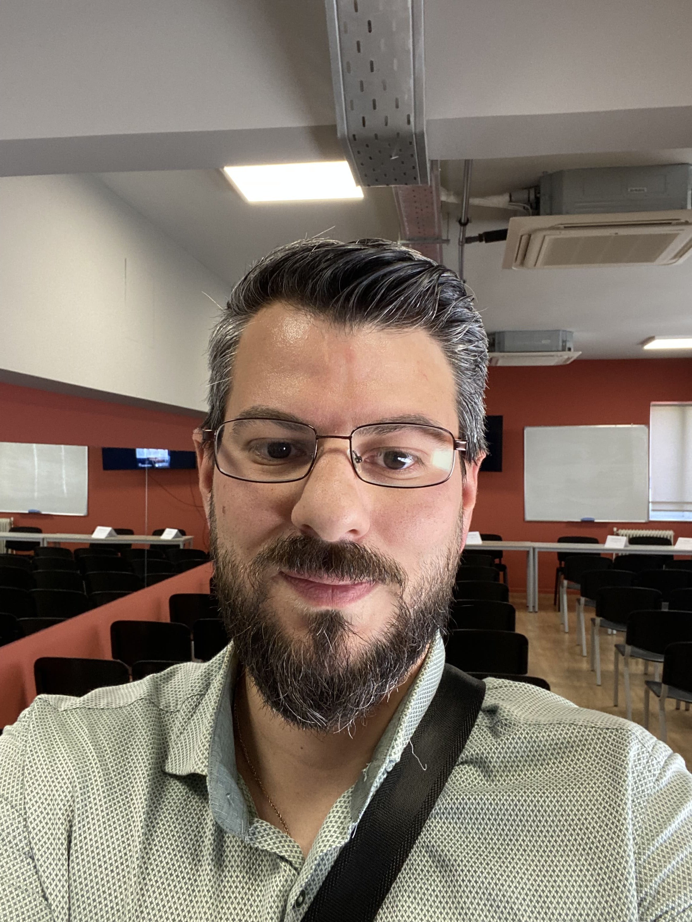 A man with glasses and a beard taking a selfie in a conference room with chairs, whiteboards, and screens in the background.