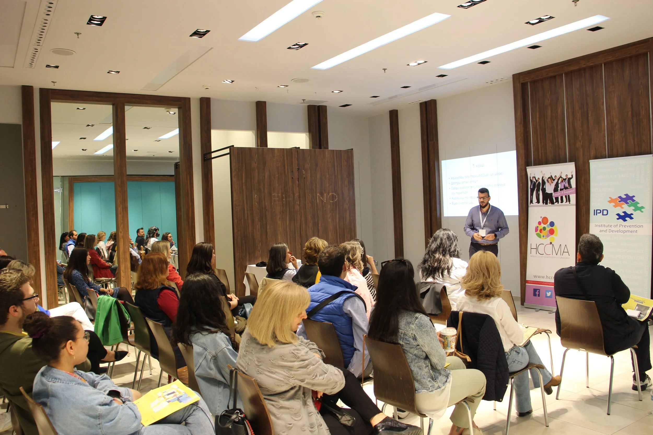 A man giving a presentation at a conference with an audience seated and listening, and banners on the side displaying logos of organizations like HCMA and IPD.