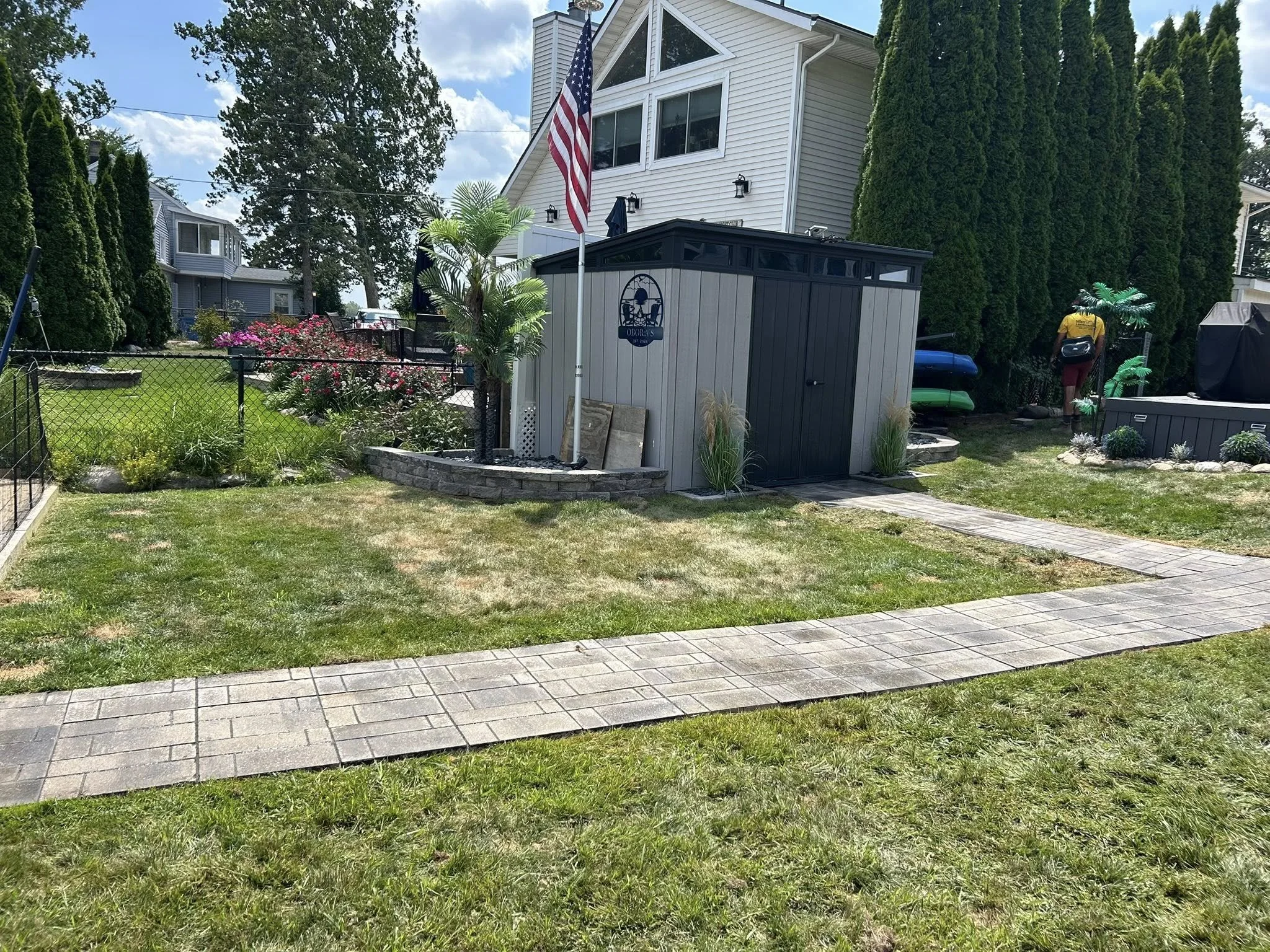 A backyard with a grassy lawn, a paved walkway, and a small shed with a flagpole featuring an American flag. There are trees and bushes, a flowerbed with roses, and a person standing near some kayaks in the background.