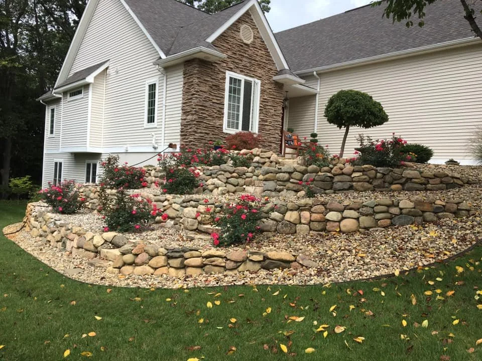 A multi-level landscaped yard with layers of rocks, pink flowering bushes, a small tree, and a porch on a white house with beige siding and a stone accent wall.