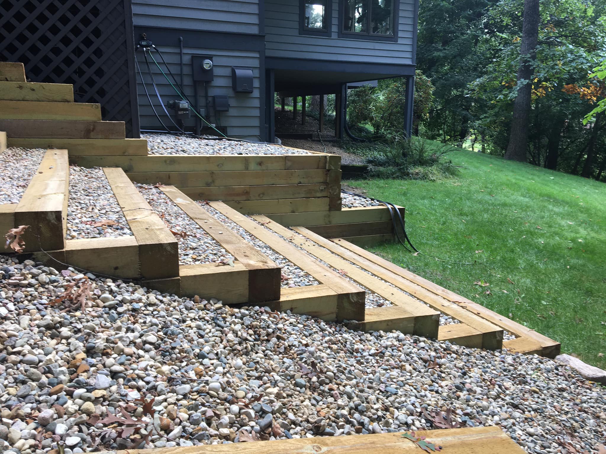 Construction of wooden stairs on a gravel pathway beside a house with a lush green yard and trees in the background.