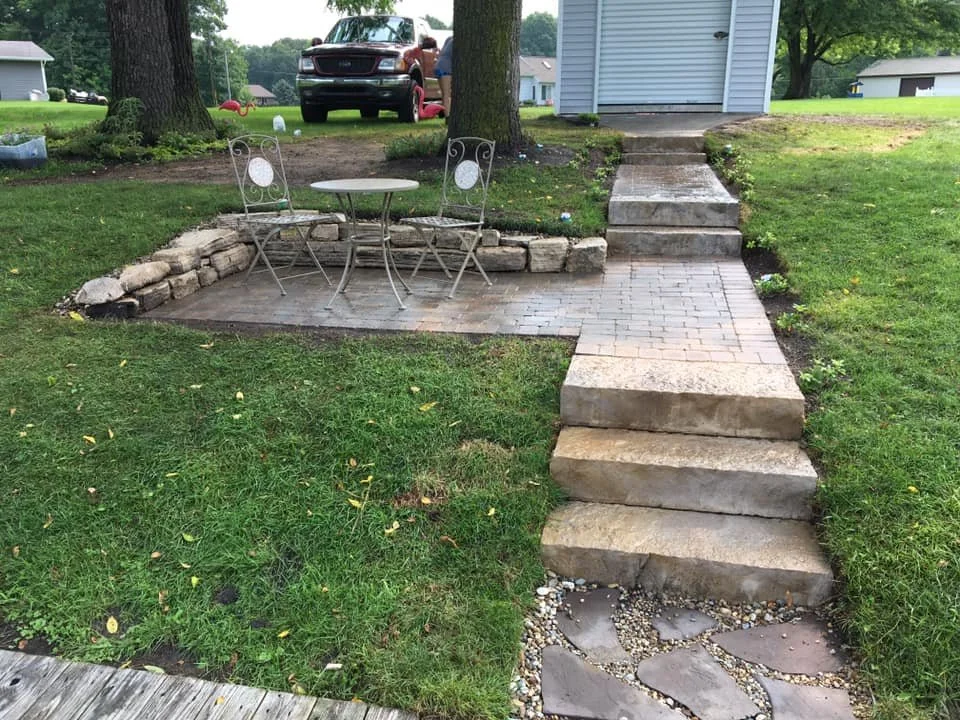 A backyard patio with a small metal table and two chairs, surrounded by grass, trees, and a stone retaining wall, with steps leading up to a house and a garage.