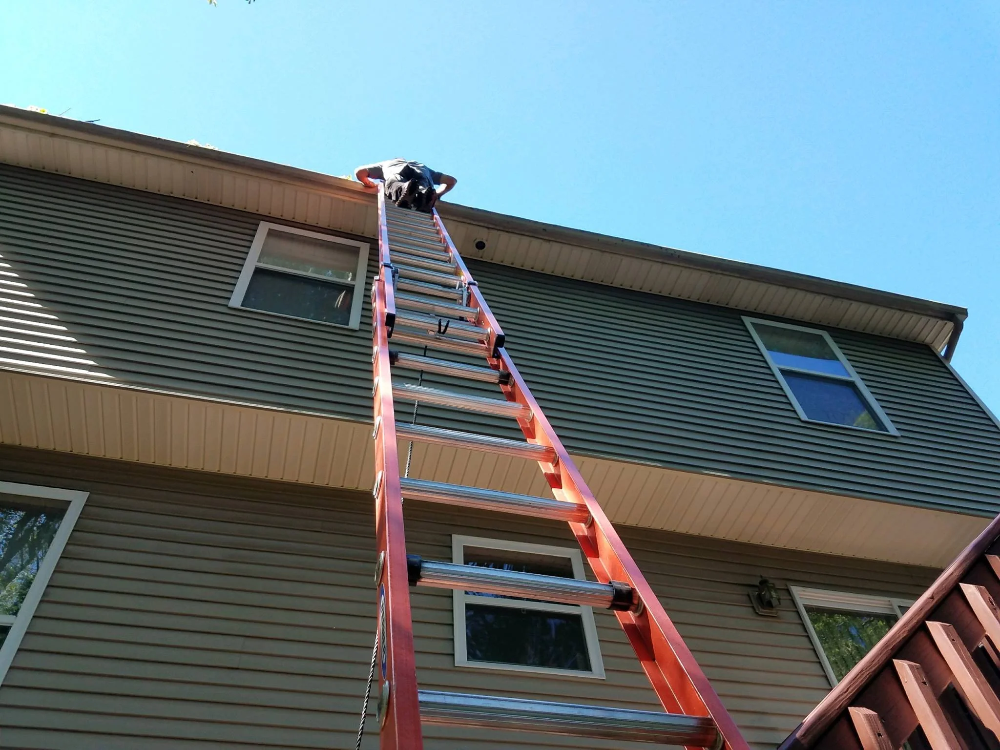 A person on a red extension ladder cleaning the roof of a house under a clear blue sky.