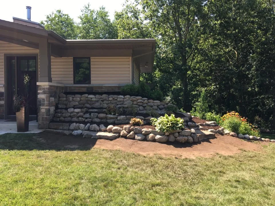 A backyard with a house, stone retaining wall, and landscaped garden beds with plants and flowers.