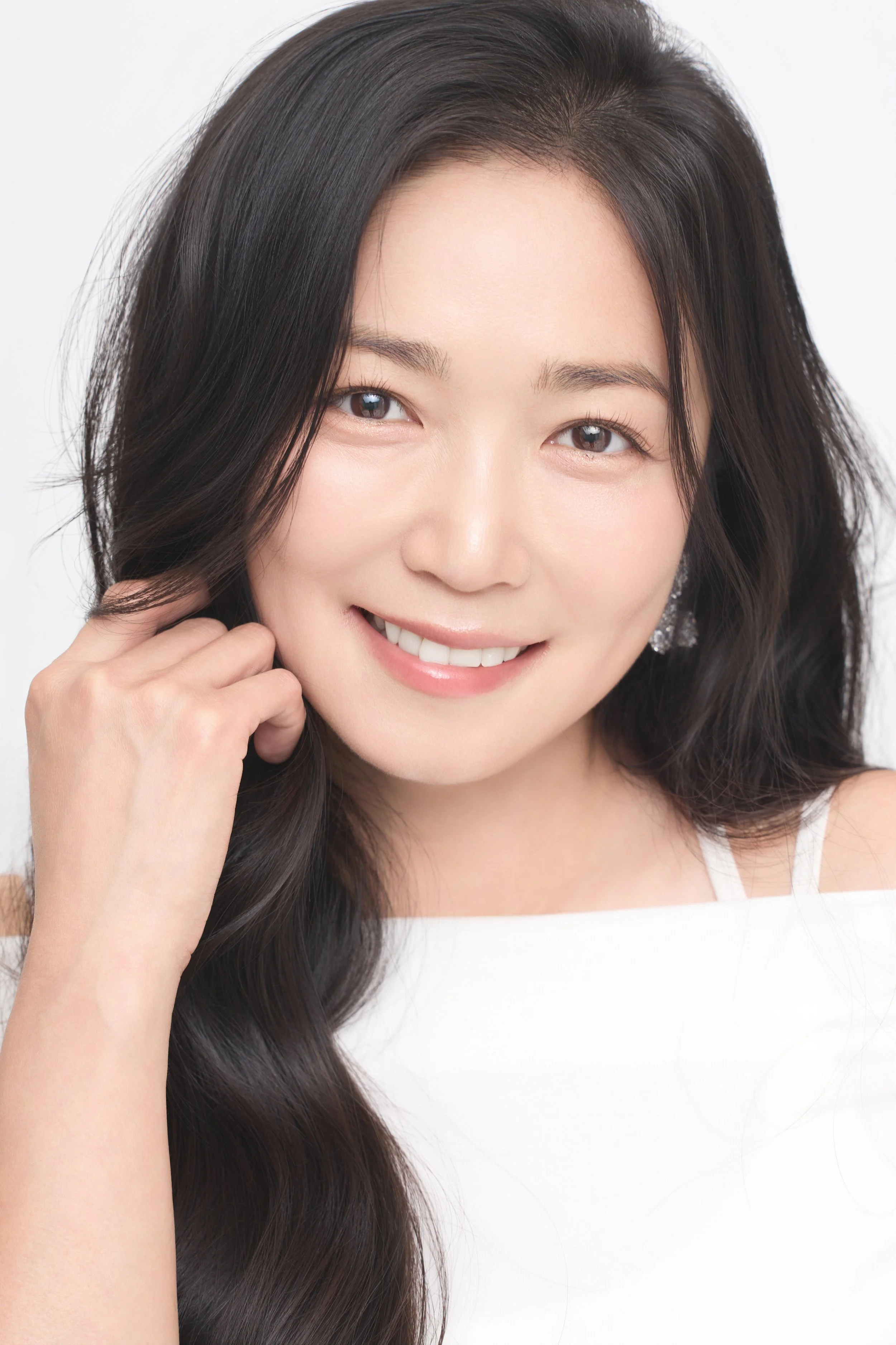 Close-up of a smiling woman Sarah Chan with long, wavy black hair, wearing earring and touching her face against a white background.