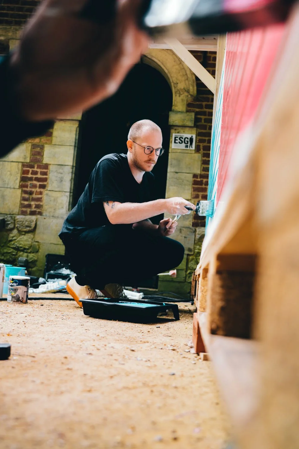 A man crouching on the floor painting a colorful design on a large surface, with various paint supplies scattered around, in a room with brick and stone walls.