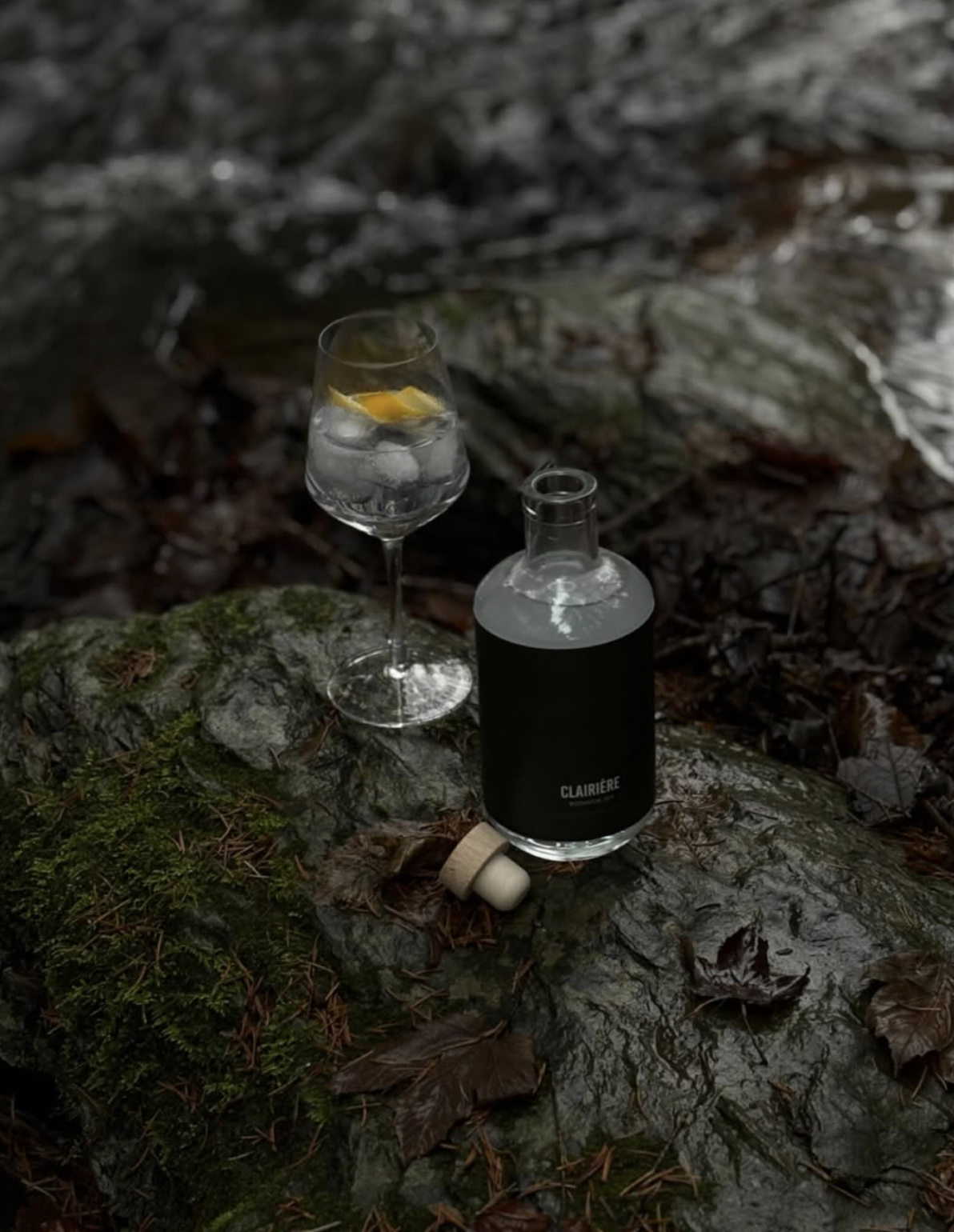 Une bouteille noire avec le mot 'Clairière' écrit dessus, un verre d'eau avec des glaçons et un citron, posé sur une roche dans un environnement forestier humide avec des feuilles mortes.