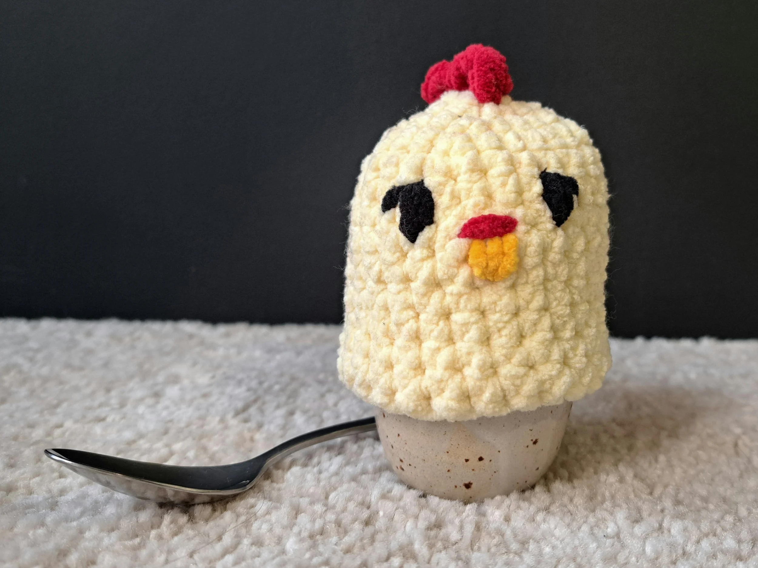 Eierwärmer "Kücken" über ein ei gestülpt, welches in einem Eierbecher steht, vor schwarzem Hintergrund