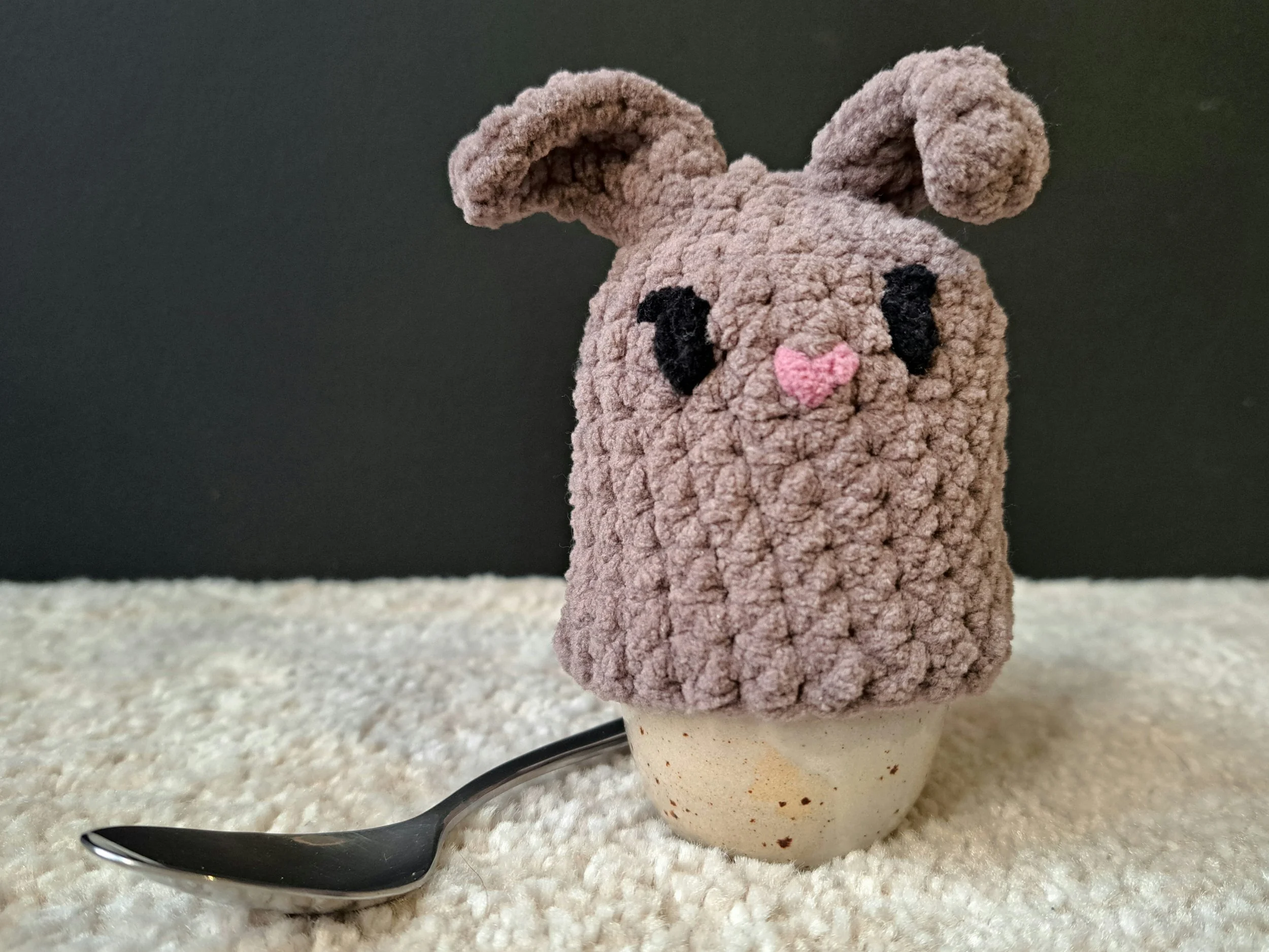 Eierwärmer "Hase dunkel" über ein ei gestülpt, welches in einem Eierbecher steht, vor schwarzem Hintergrund