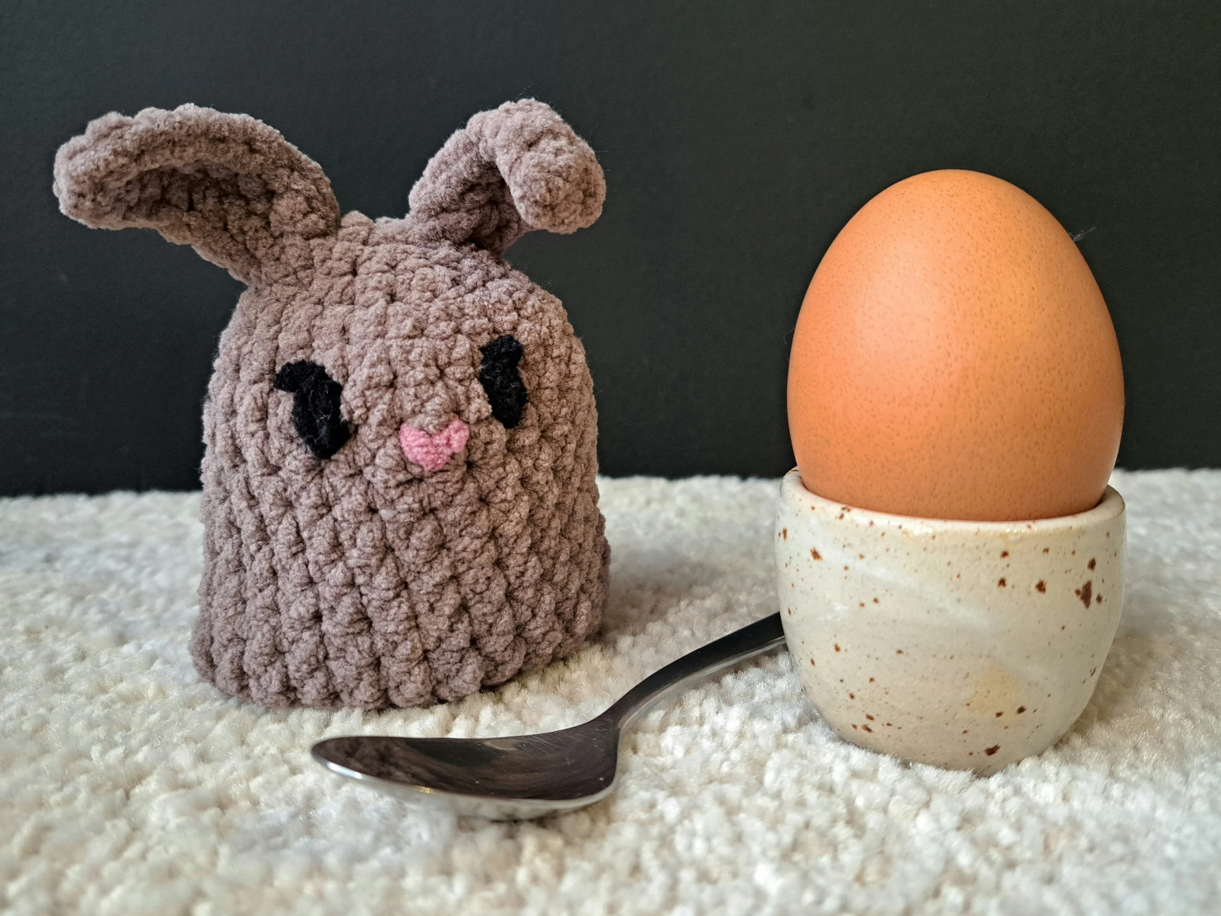 Eierwärmer "Hase dunkel" steht auf dem Boden, rechts ist ein ei in einem Eierbecher von Töpferhof Ulrike Rost zusehen