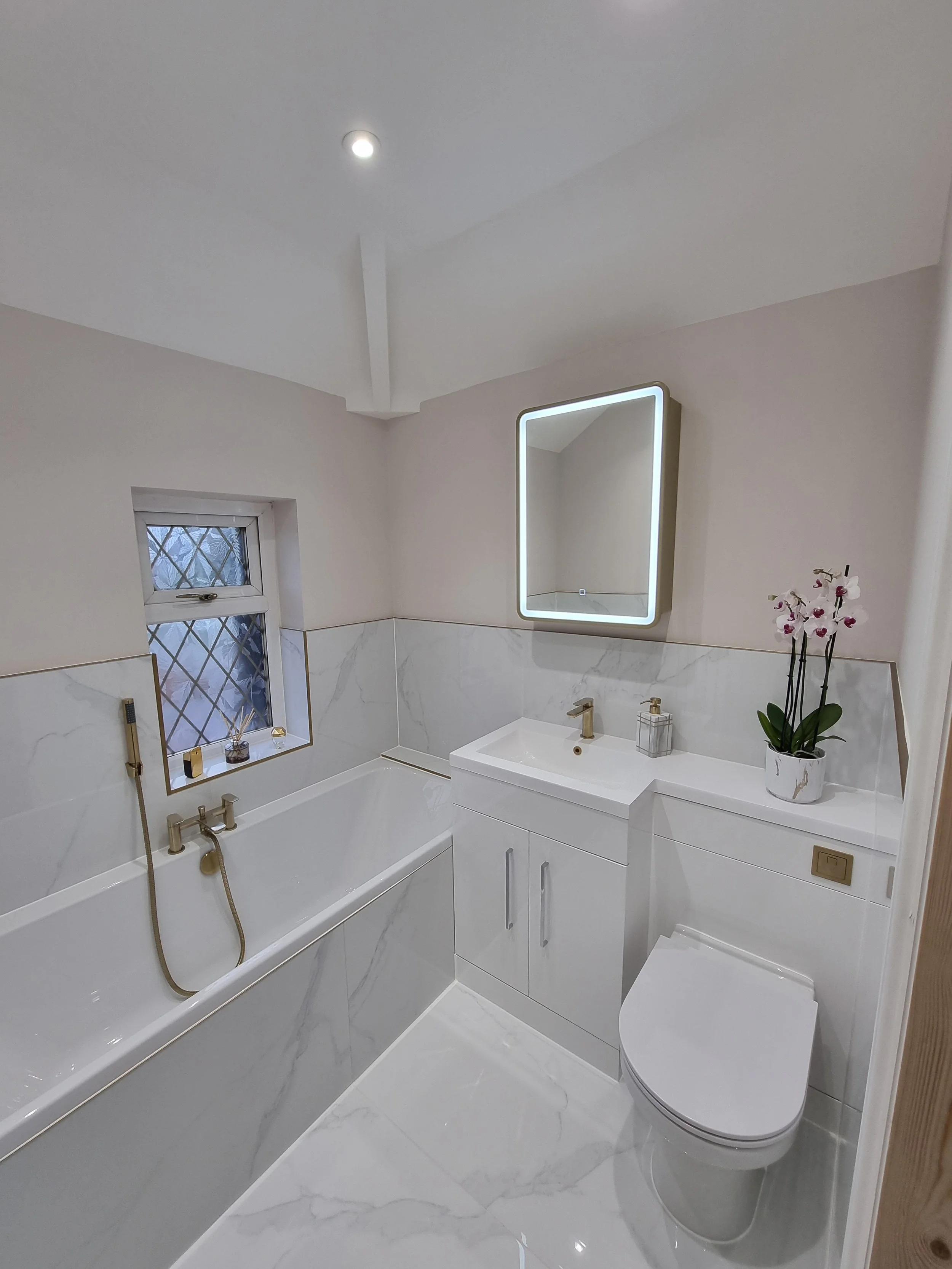 Modern small bathroom with white marble floor and walls, a bathtub with a gold faucet, a small window with a decorative grid, a white vanity with a built-in sink, a illuminated mirror, a potted orchid, and a wall-mounted toilet.