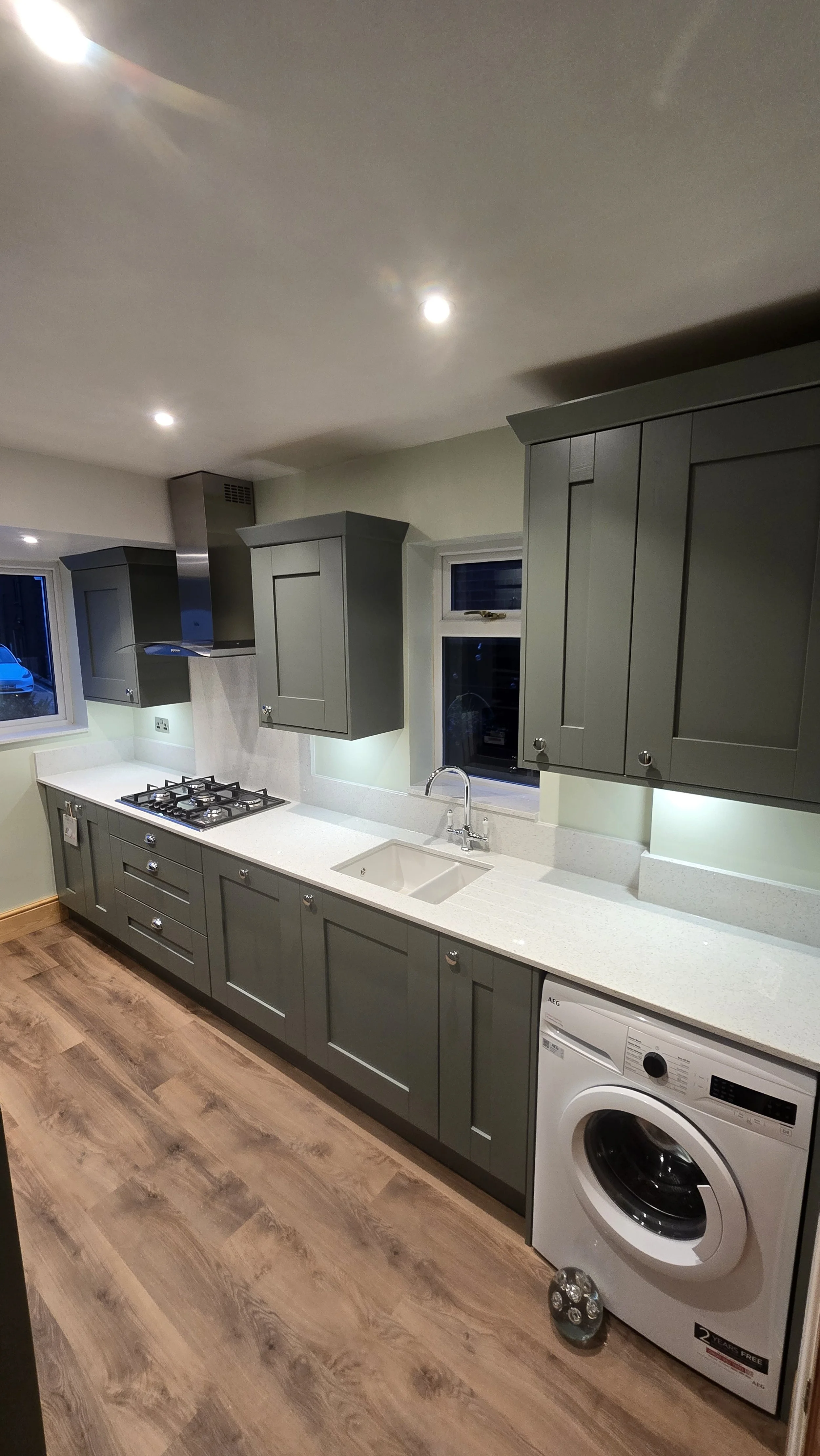 Modern kitchen with gray cabinets, white countertop, gas stove, sink, window, and washing machine, wood flooring.