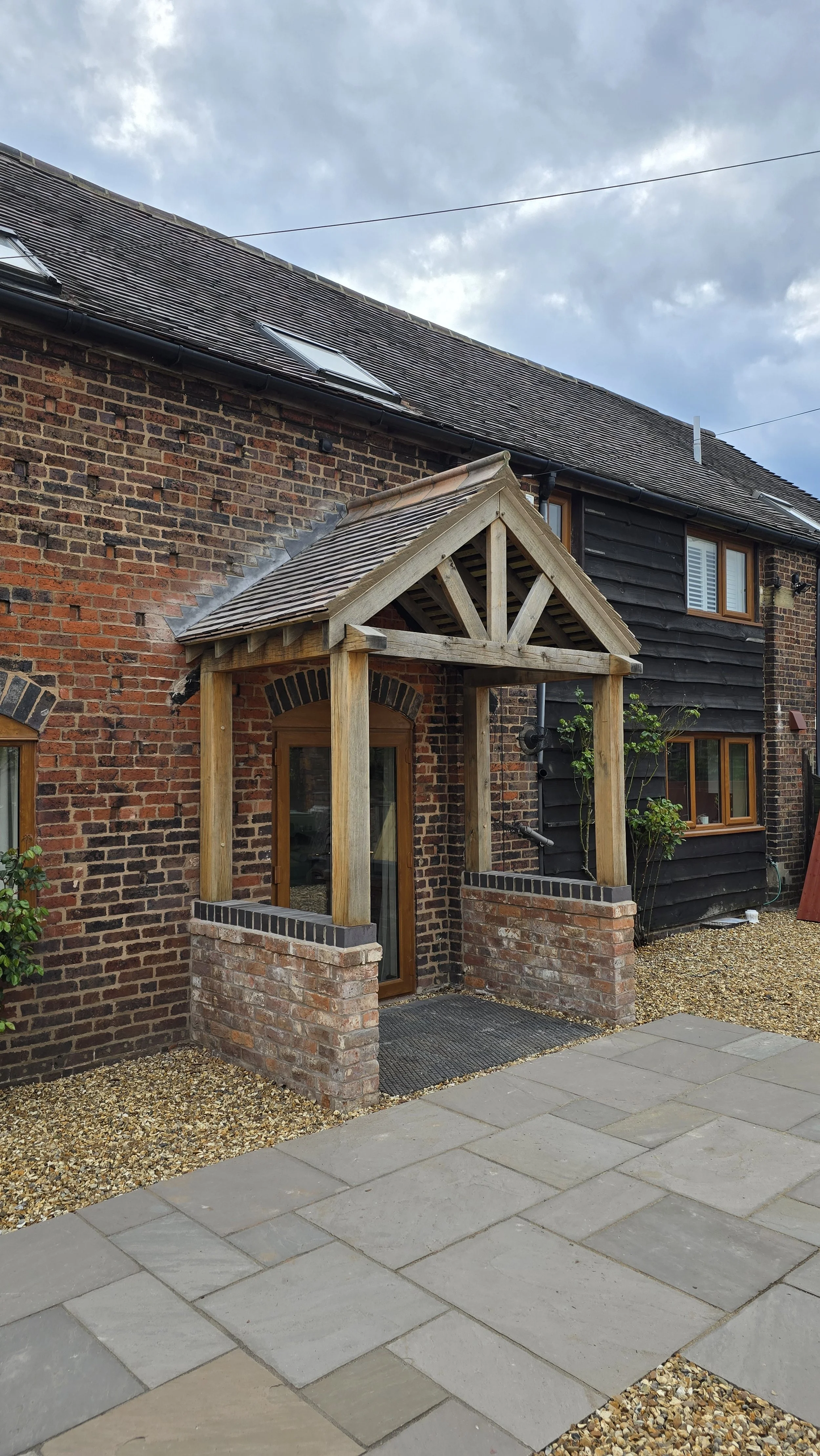 A brick house with a small wooden porch structure over a door, featuring wooden beams and a slanted roof. There are windows with wooden frames, and the house has a mix of red brick and black wooden siding. The yard has paved pathways and gravel areas.