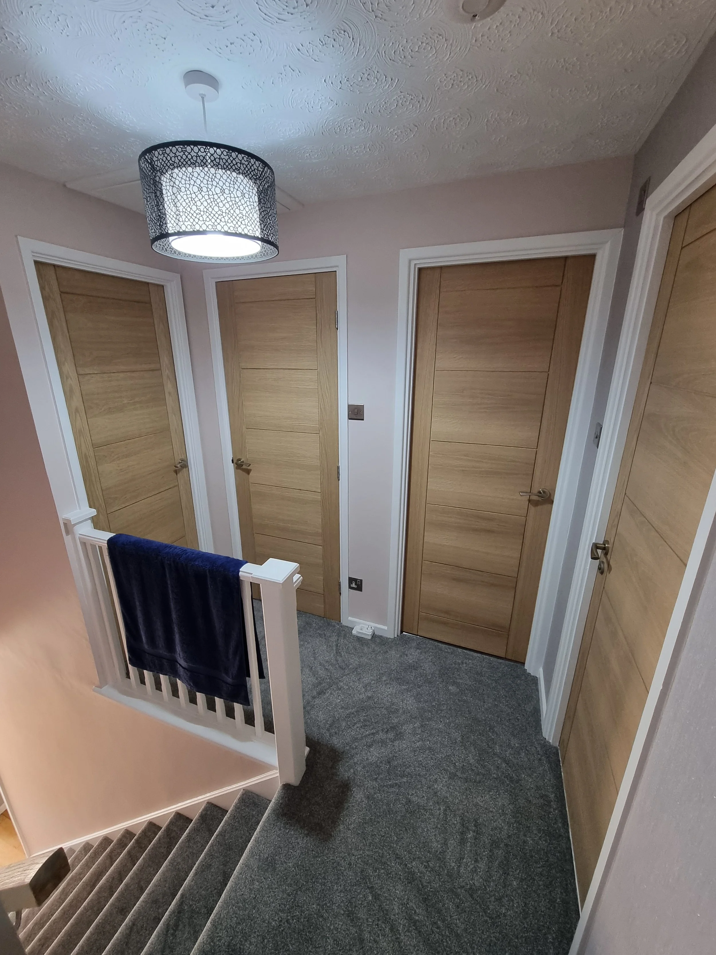 A small hallway with four wooden doors, a gray carpeted floor, a staircase with a navy towel, beige walls, a ceiling light fixture, and white trim.