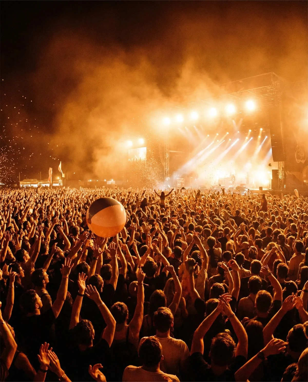 Crowd at a concert or music festival with hands raised, a large beach ball, and bright stage lights with smoke or fog effects.