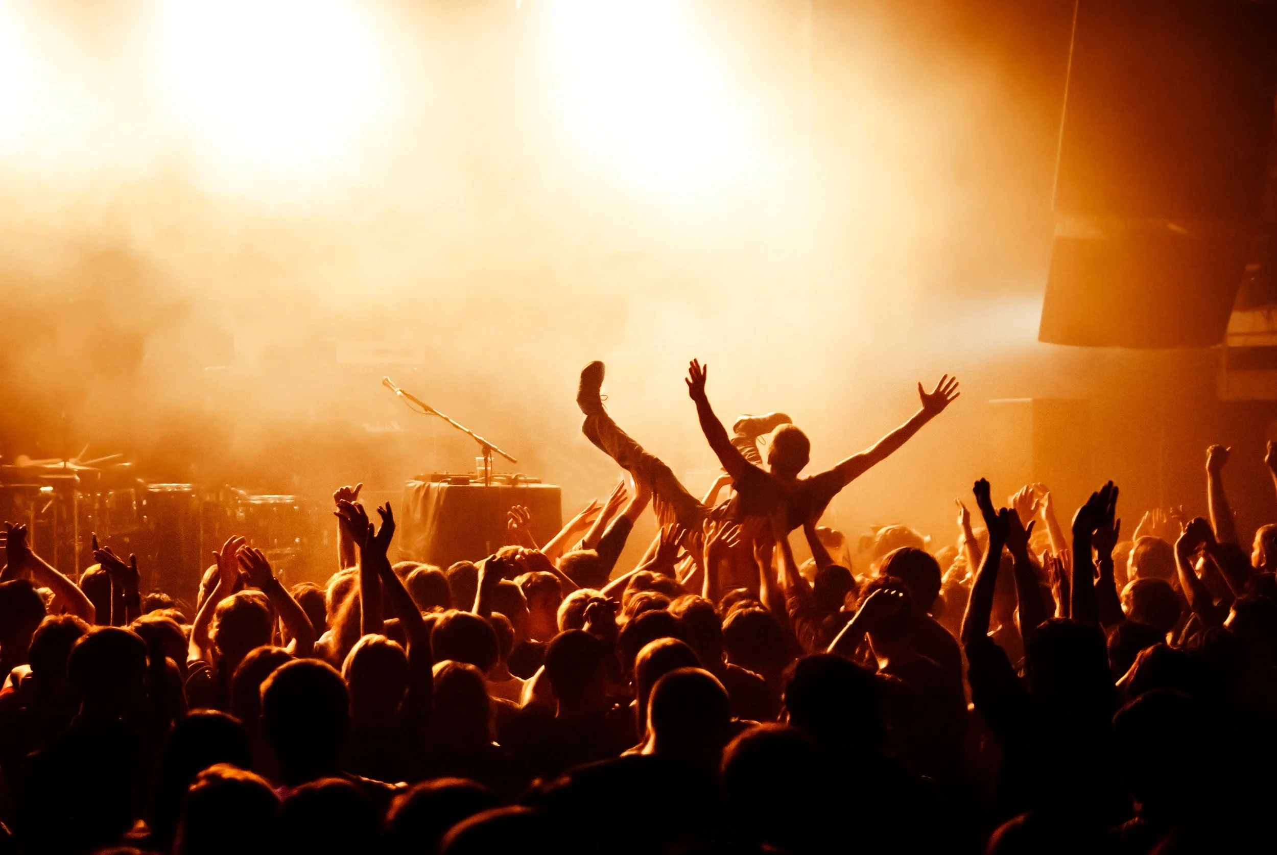 Crowd of people at a concert or music event with hands raised, bright stage lights shining in the background.