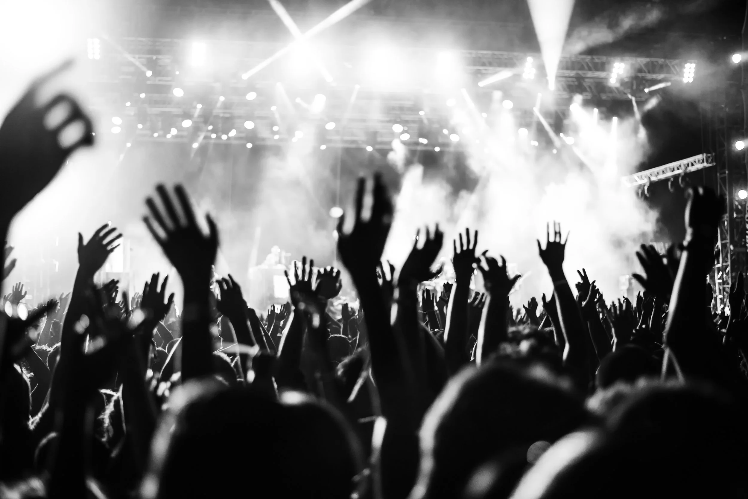 Black and white photo of a concert crowd with hands raised, stage lights and smoke creating an energetic atmosphere.