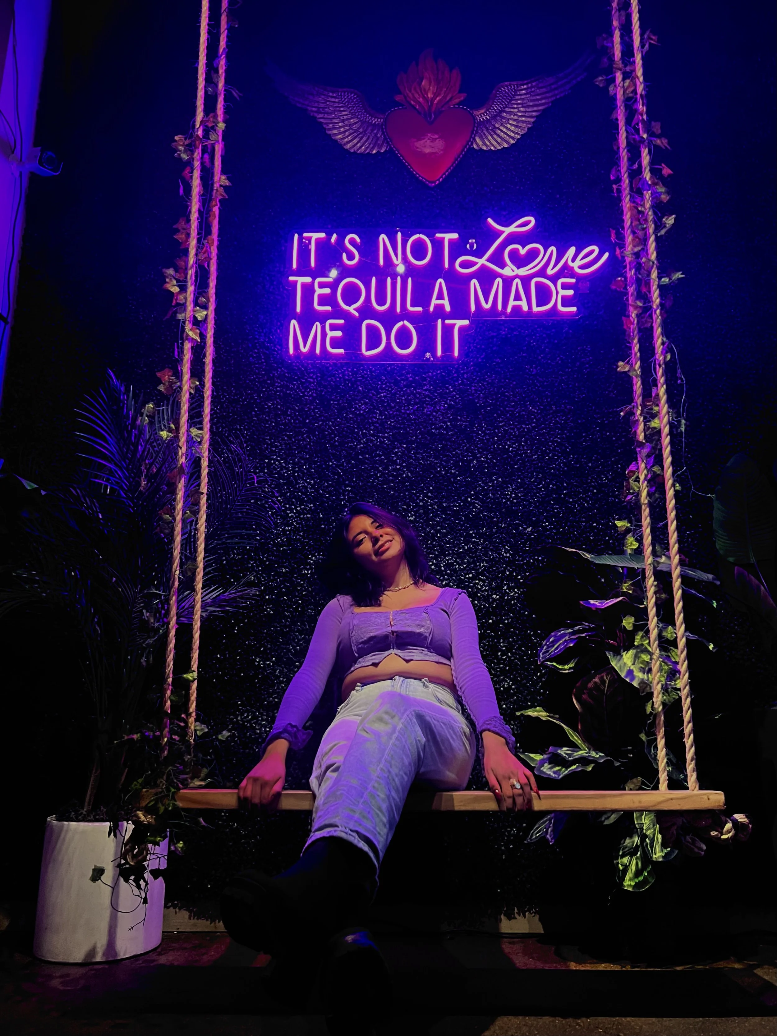 Woman sitting on a wooden swing in front of a black textured wall illuminated by purple neon lights, with a neon sign that says 'It's not love tequila made me do it' and a decorated heart with wings above.