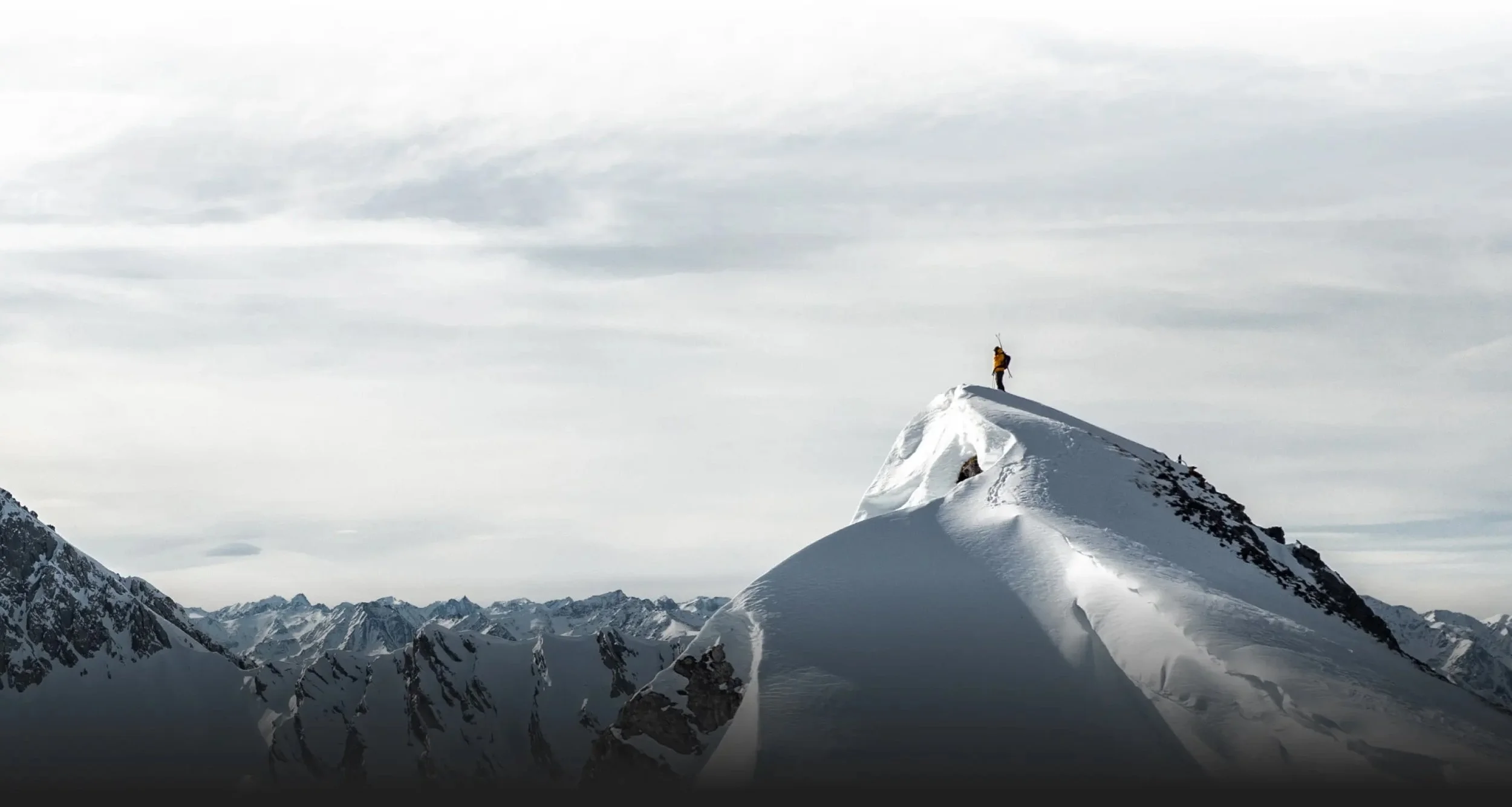 Suunto. A person wearing yellow jacket standing at the summit of a snow-covered mountain peak, with a vast range of snowy mountains in the background on a cloudy day.