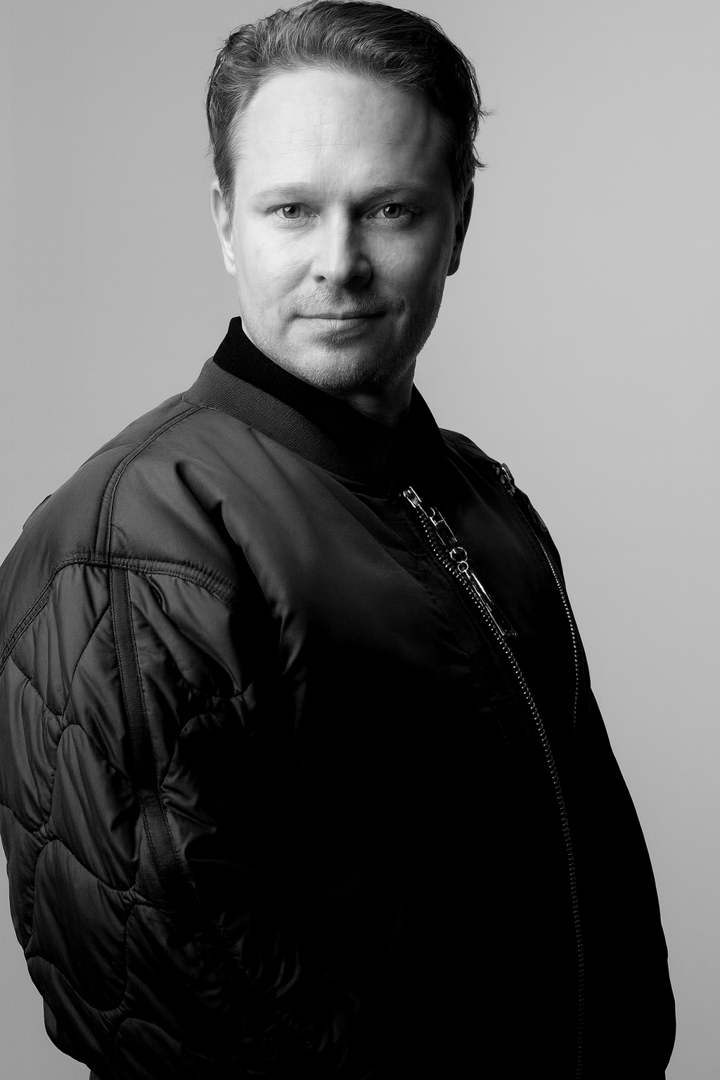 Black and white portrait of a man with short hair, wearing a leather bomber jacket, looking at the camera.