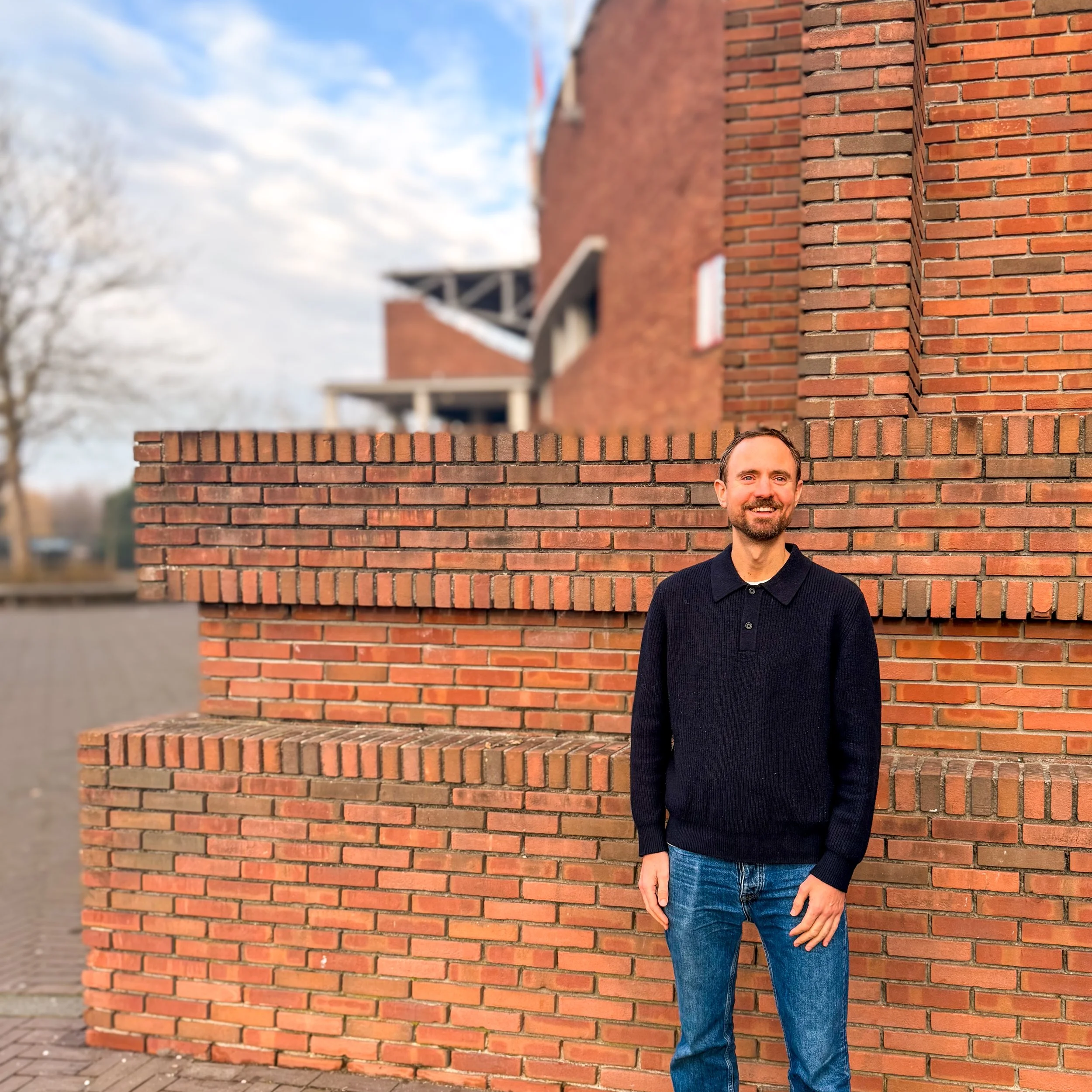 A man with a beard and short hair wearing a black sweater and jeans, smiling, standing in front of a brick wall.
