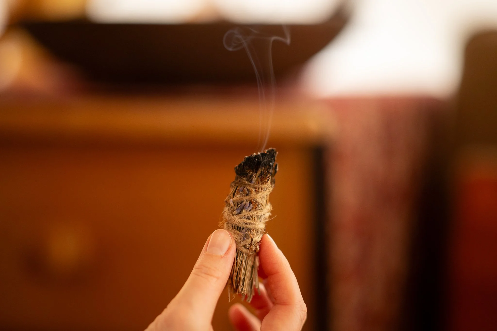 A person's hand holding a smoldering bundle of herbs or incense with thin smoke rising from it.