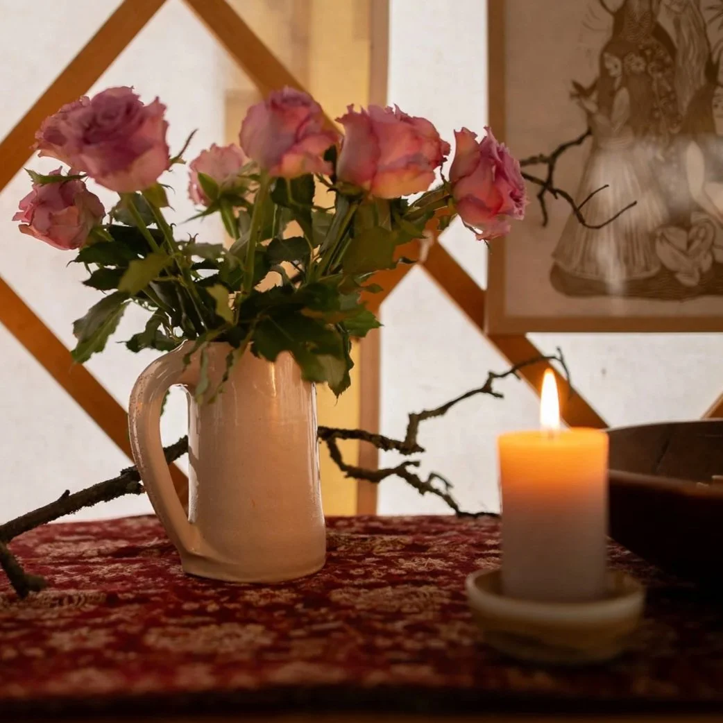 Pink roses in a white pitcher on a table with a lit candle and decorative branches in the background.