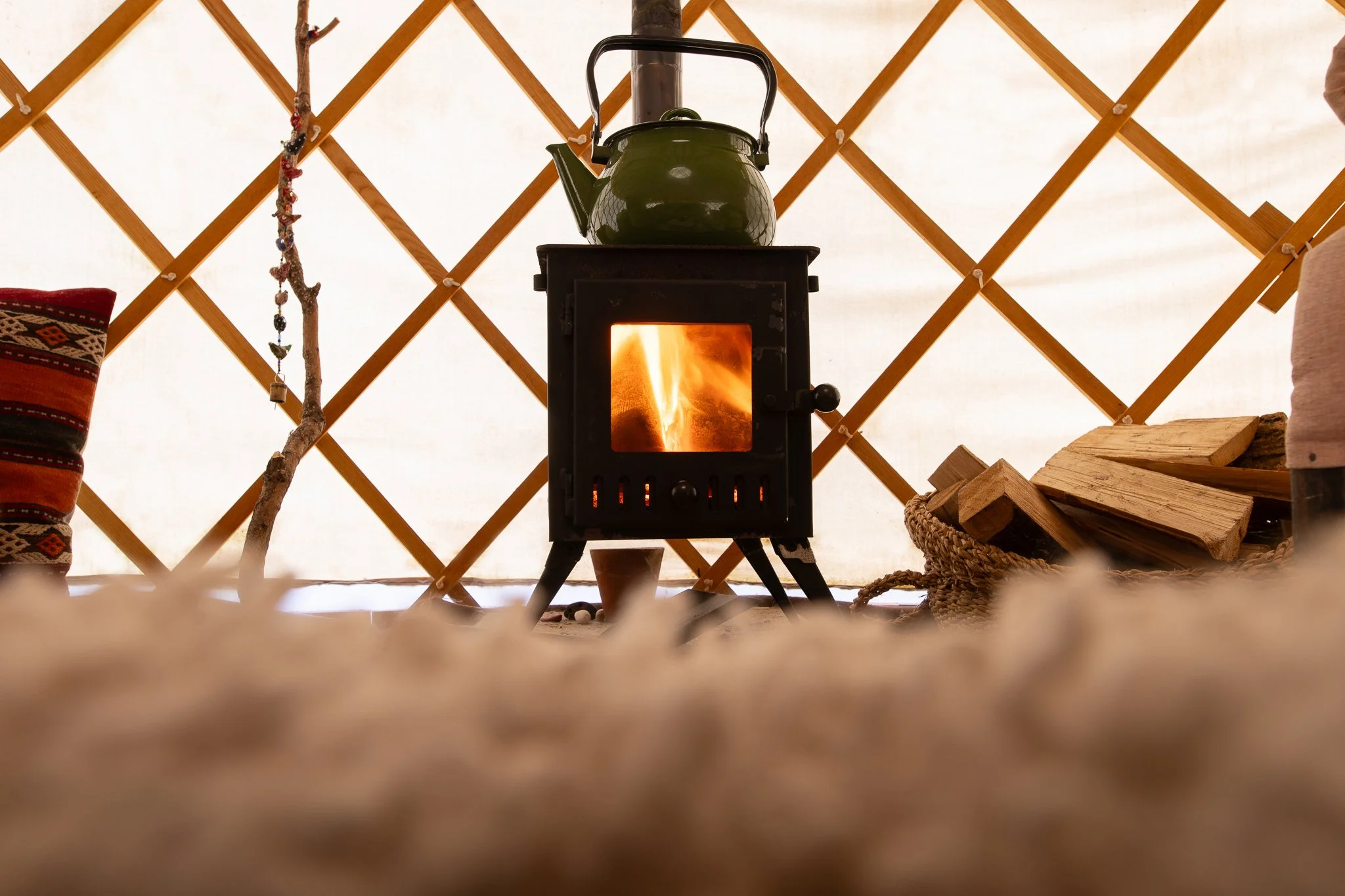 A wood stove with a glass door showing a fire inside, with a green kettle on top, inside a yurt with a lattice wall and some firewood and decorations.