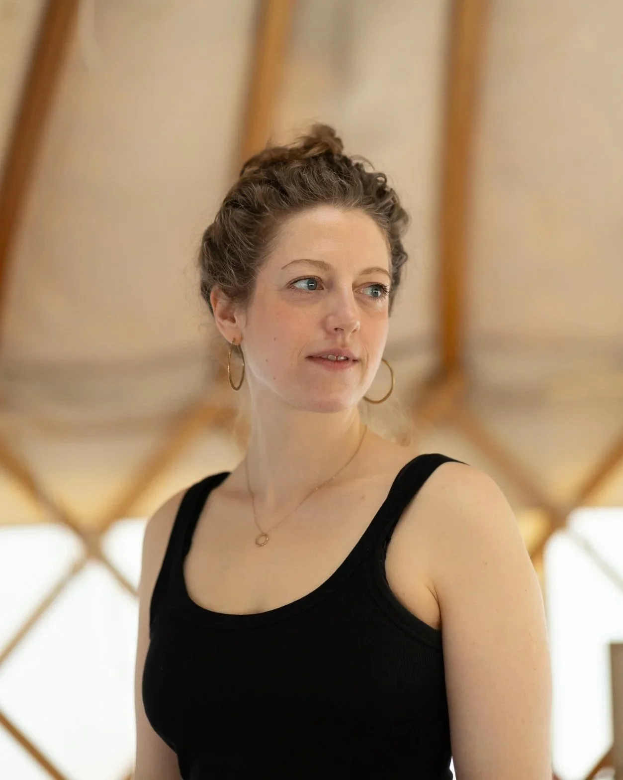 A woman with curly hair tied up, wearing gold hoop earrings, a delicate necklace, and a black tank top, standing indoors in front of a wooden ceiling structure.