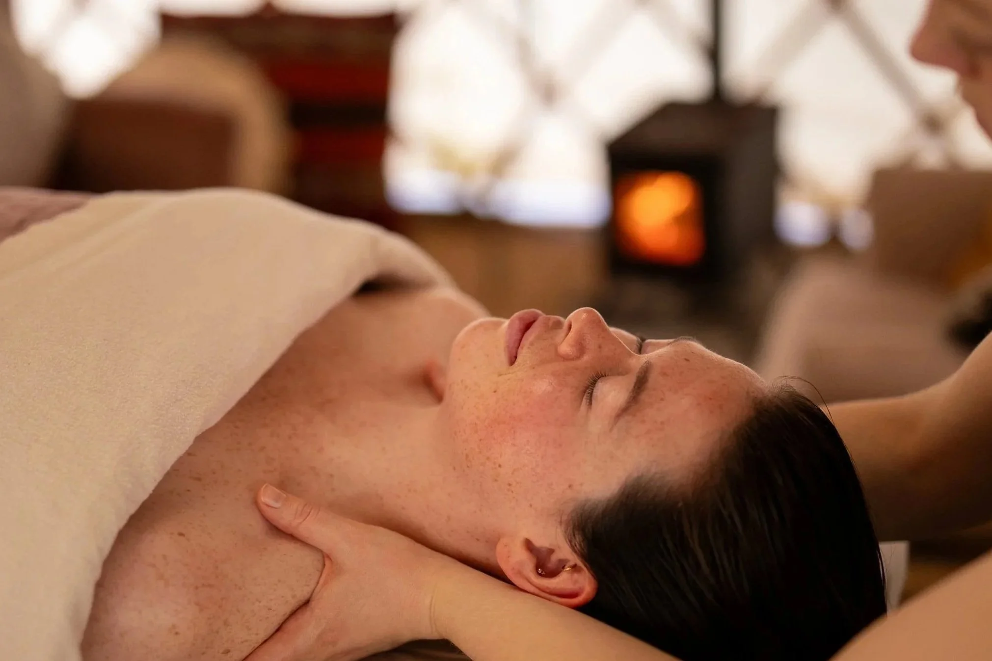 A woman lies on her side with eyes closed, enjoying a massage in a peaceful setting. She has dark hair, light skin with freckles, and is wearing earrings. The background features warm lighting and a cozy ambiance.