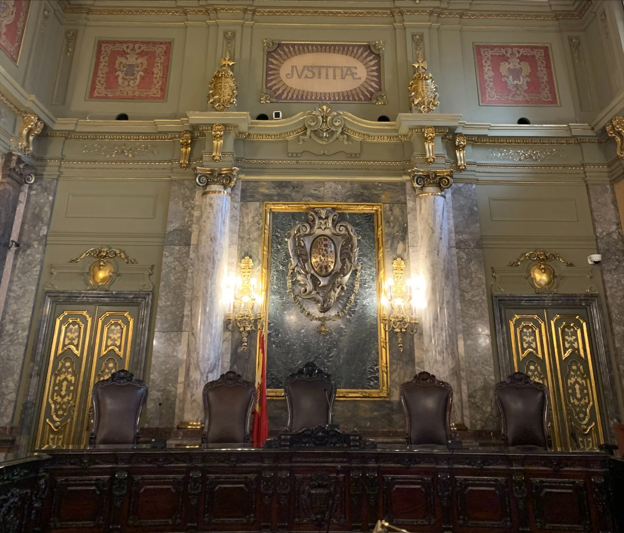 Sala de tribunal con cinco sillas de cuero y madera oscura, decorada con columnas de mármol, candelabros y un escudo enmarcado en la pared detrás del estrado. En la parte superior, la palabra 'JUSTICIA' en letras mayúsculas.