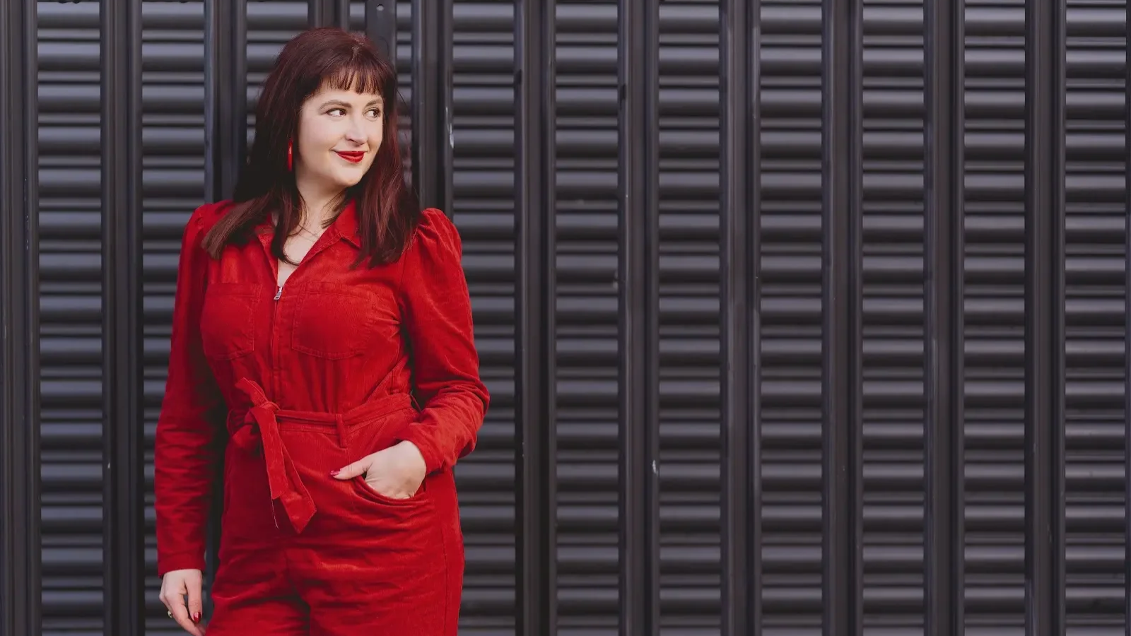 Elin Edwards, wearing a red jumpsuit and red lipstick, standing in front of a black metal fence, posing and smiling.