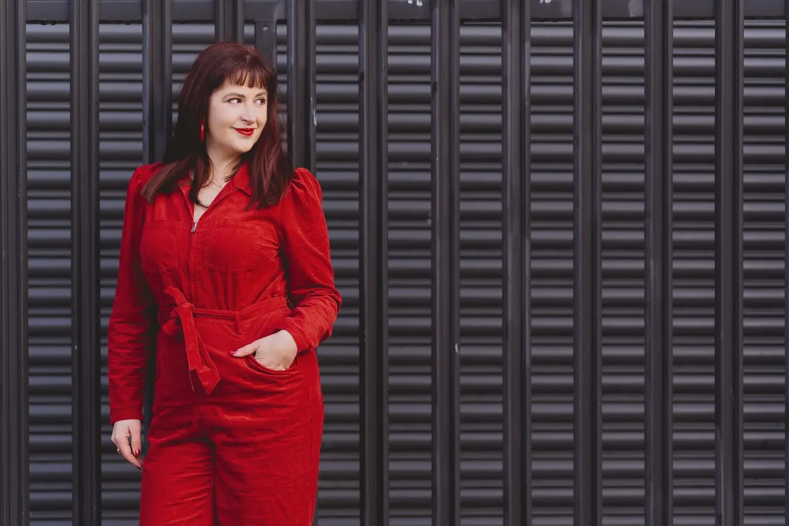 Elin wearing a red jumpsuit with a tie at the waist, standing in front of a black metal fence and looking off to the side.