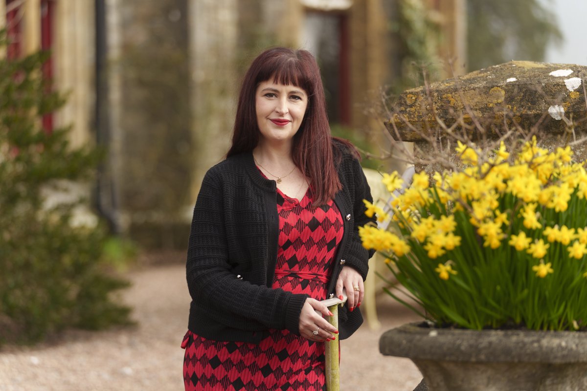 Elin in a red and black patterned dress standing outdoors next to a large pot of yellow daffodils.