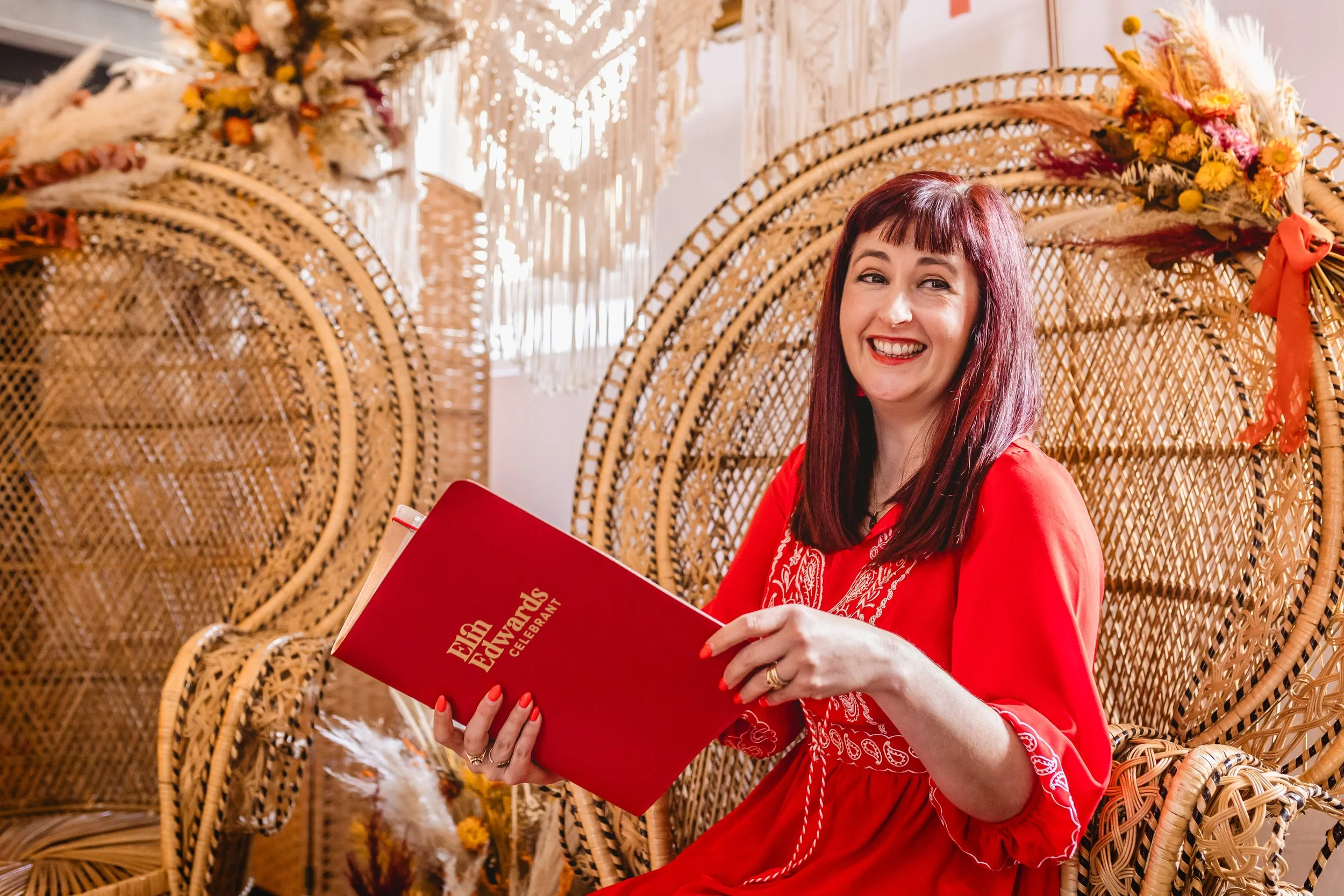 Elin in a red dress is sitting on a wicker chair, smiling, and holding a red celebrant folder with "Elin Edwards Celebrant" embossed. The background features a decorative, boho-style arrangement with flowers.