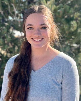 A young woman with long brown hair smiling outdoors, wearing a light gray T-shirt with trees in the background.
