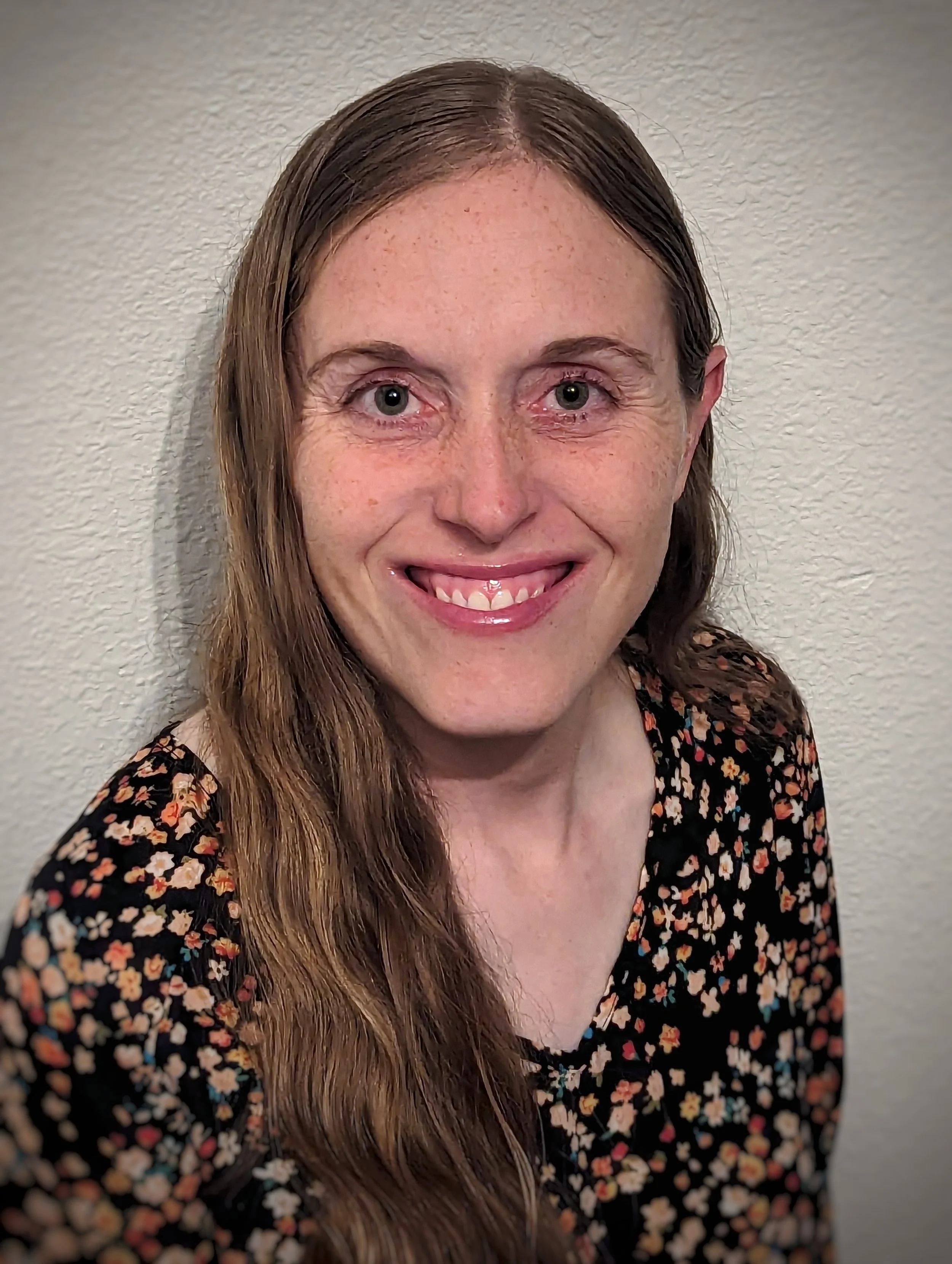A woman with long brown hair, blue eyes, and freckles smiling and sticking out her tongue, wearing a black floral-patterned top, standing against a white textured wall.